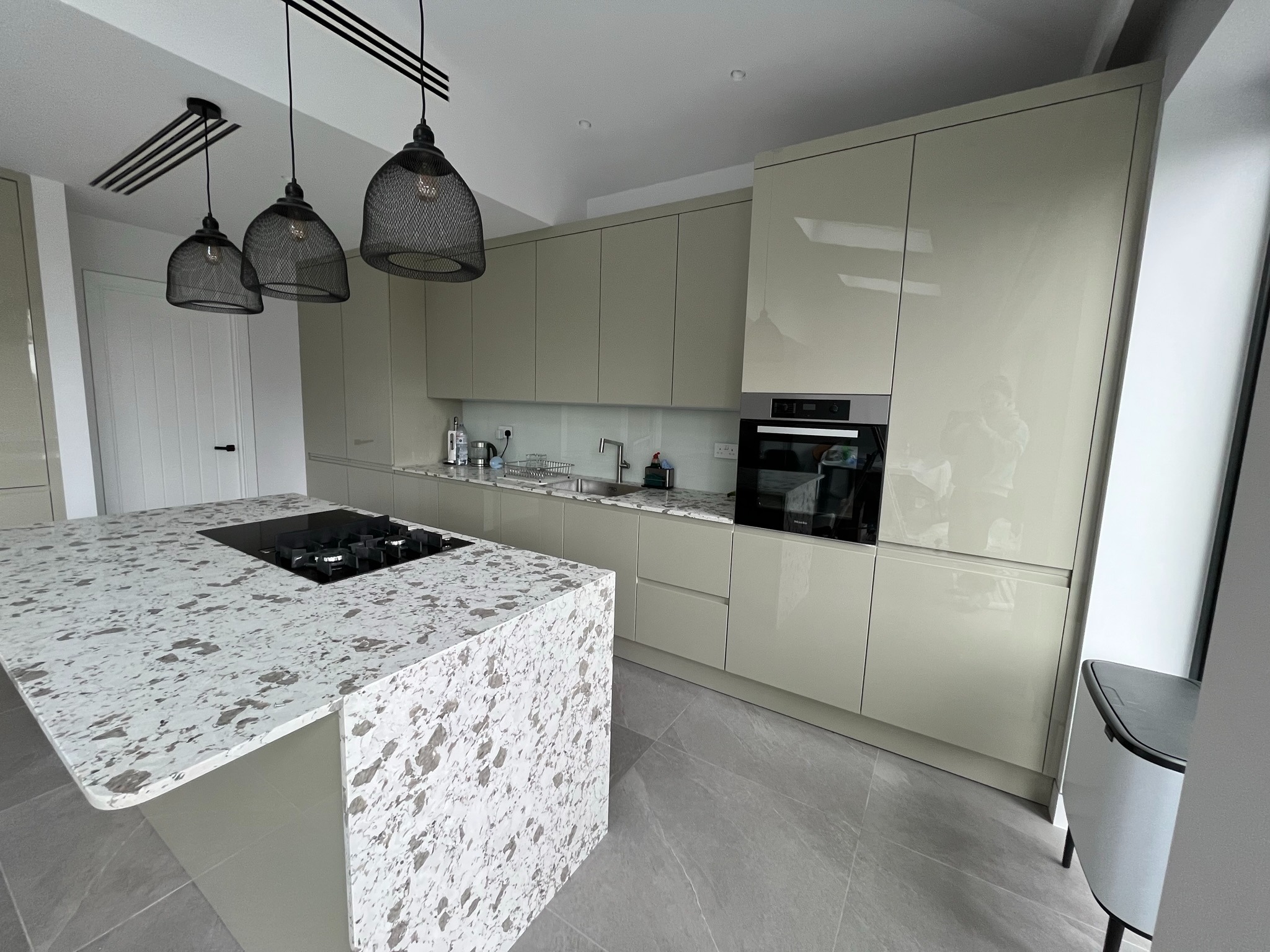 Beżowa kuchnia z wyspą i blatem w plamki, oświetlona trzema lampami z metalowej siatki. Błyszczące fronty szafek, zabudowany piekarnik, płyta grzewcza na wyspie.