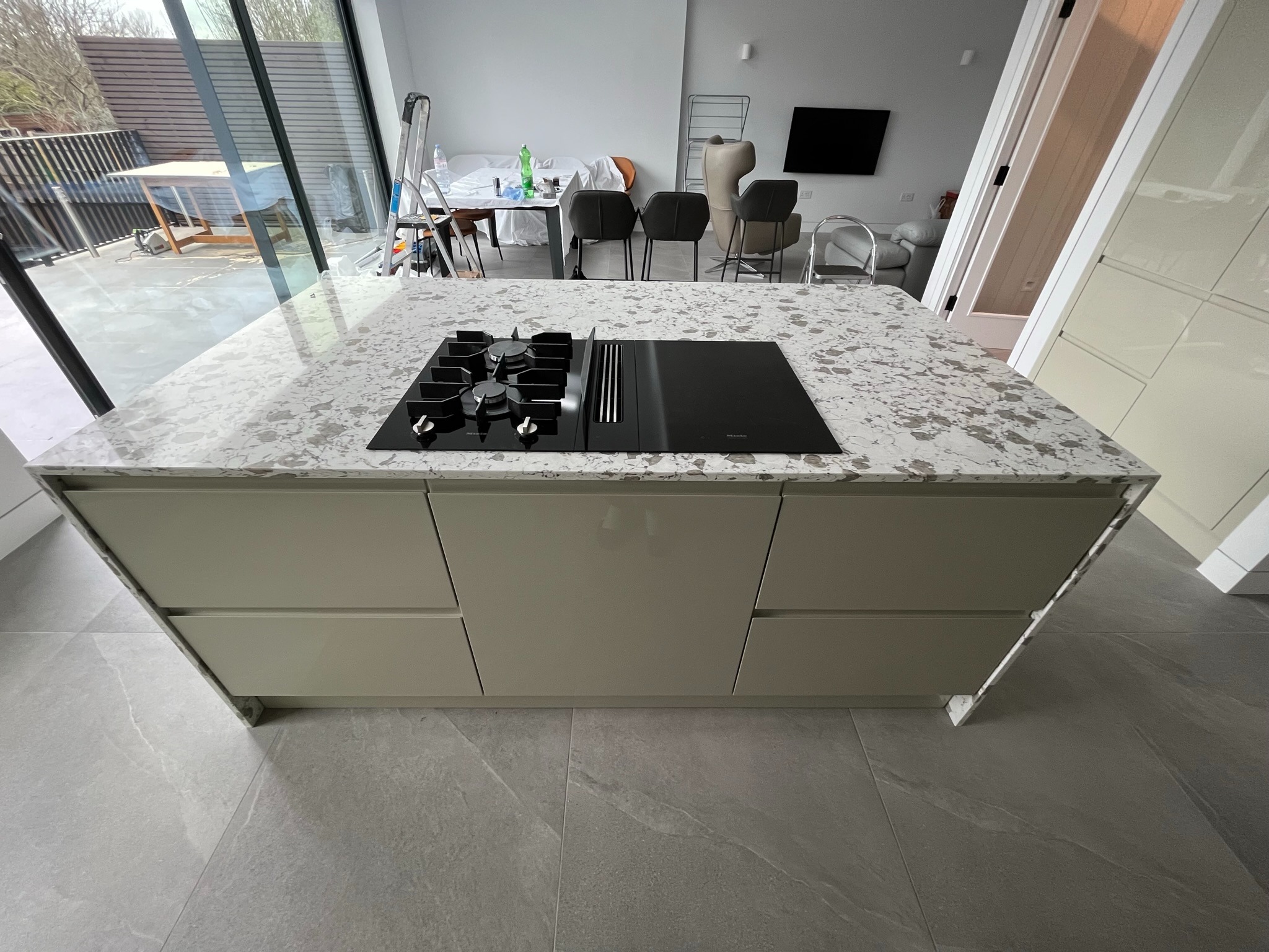 Wyspa kuchenna z szarymi szafkami i blatem w plamki, z wbudowaną płytą grzewczą Miele. W tle okno i elementy wyposażenia wnętrza.