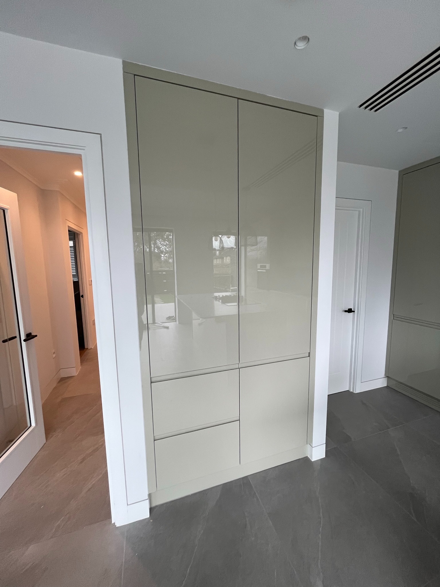 Beżowa szafa wnękowa z połyskiem, wkomponowana w jasne wnętrze. Widoczne drzwi po lewej i prawej stronie, szara podłoga, minimalistyczny design.