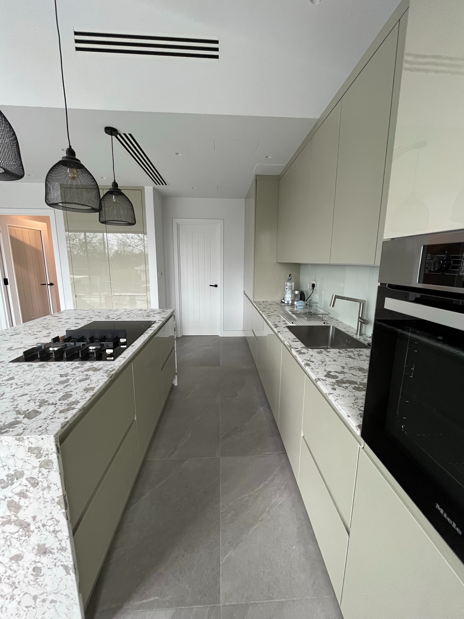 Nowoczesna kuchnia z wyspą i blatem w plamki, oświetlona wiszącymi lampami z metalowej siatki. Widoczny piekarnik Miele i szare płytki podłogowe.