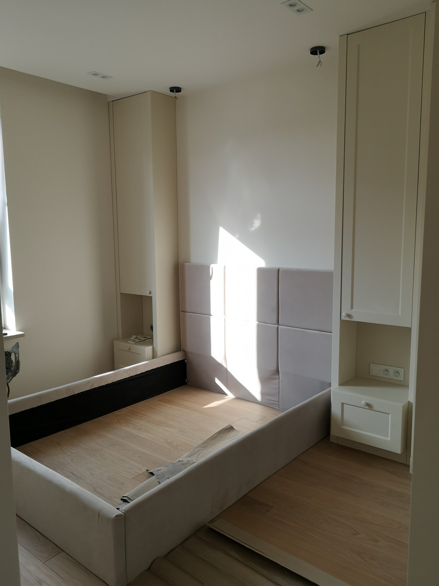 Rama łóżka tapicerowanego z wezgłowiem z paneli, wkomponowana między dwie wysokie szafy z wbudowanymi szafkami nocnymi, w jasnym wnętrzu.