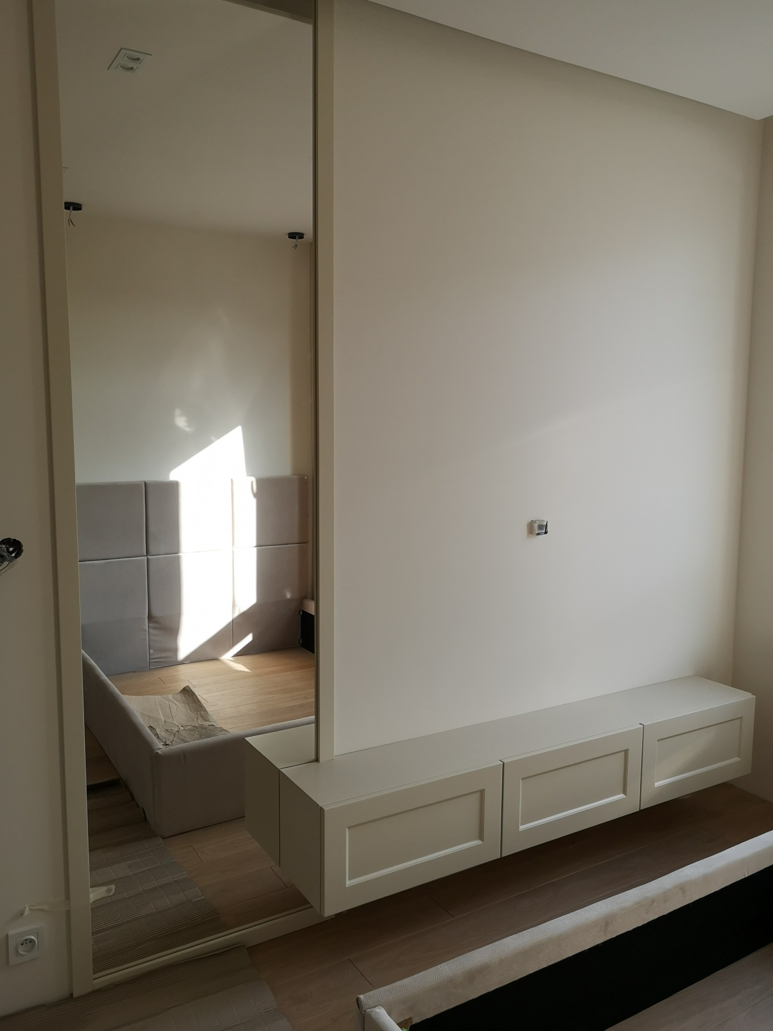 Nowoczesna szafa wnękowa z lustrem i wisząca szafka RTV w jasnej sypialni. Widoczne detale wykończenia i delikatne oświetlenie.