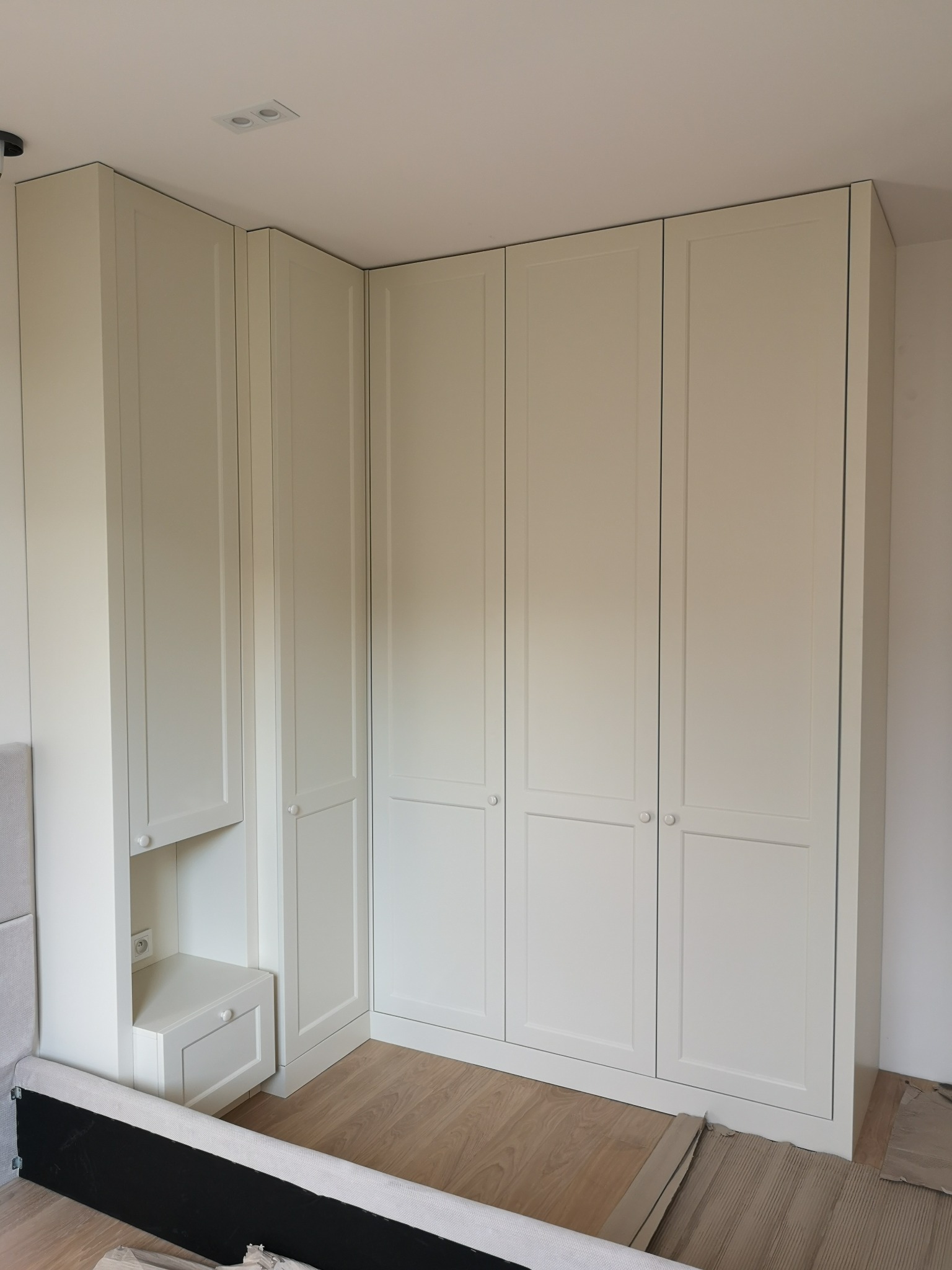 Jasna, narożna szafa wnękowa z frezowanymi frontami, wbudowana w sypialni, zintegrowana z szafką nocną z gniazdkiem elektrycznym. Ujęcie z dołu, widoczna podłoga.