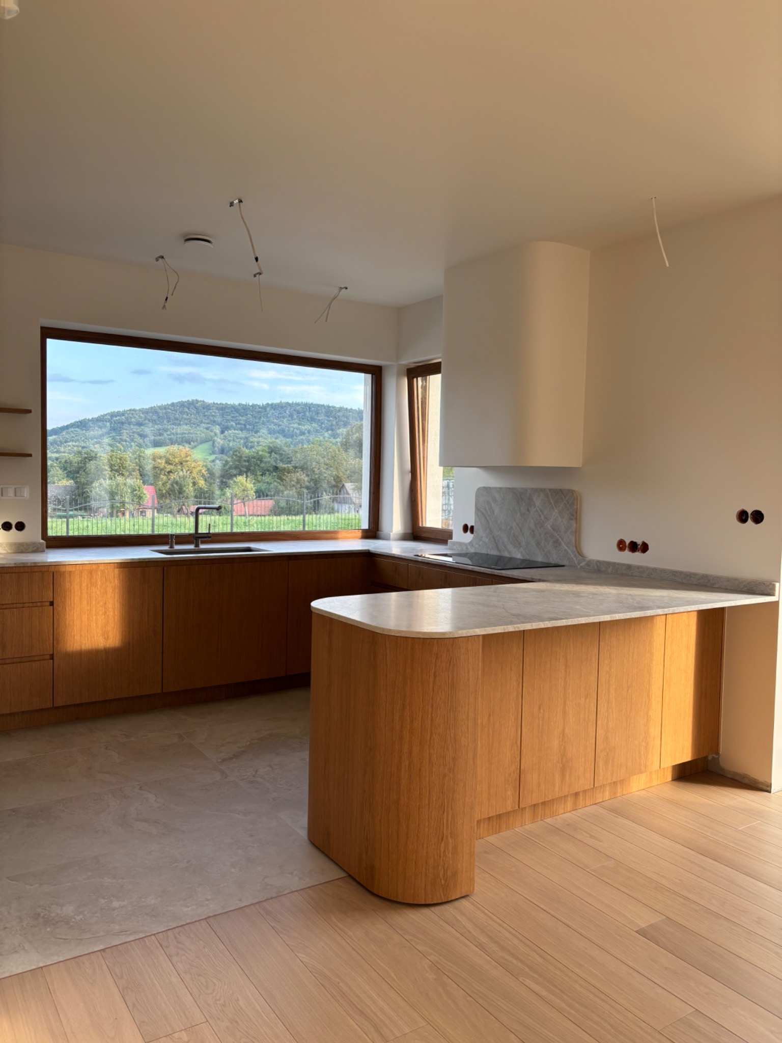 Jasna, nowoczesna kuchnia z drewnianymi frontami i marmurowym blatem. Duże okno z widokiem na góry. Elementy wykończeniowe w trakcie montażu.