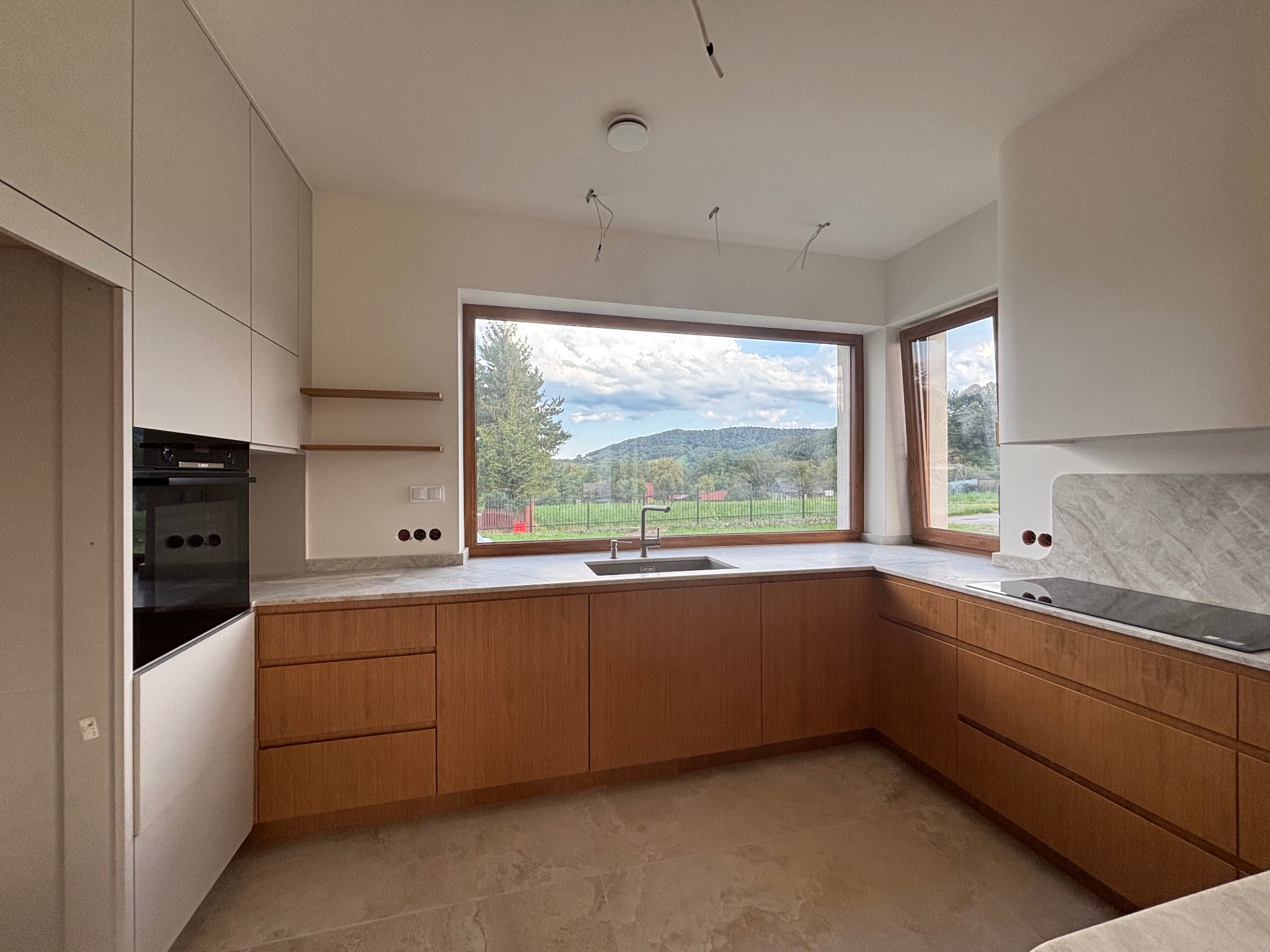 Nowoczesna kuchnia z drewnianymi szafkami i marmurowym blatem w Wadowicach. Duże okno z widokiem na zieleń. Minimalistyczny design.