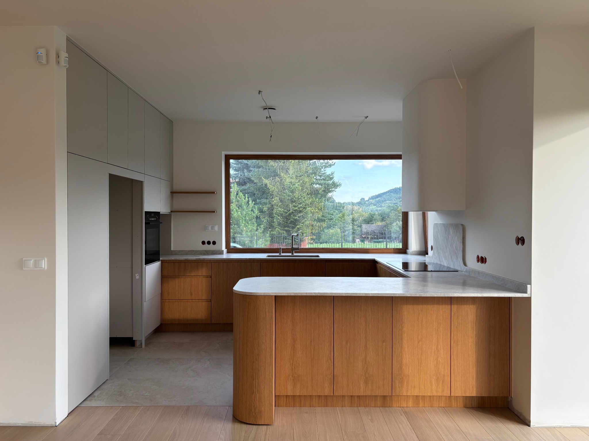 Nowoczesna kuchnia z wyspą i drewnianymi frontami. Marmurowy blat, duże okno z widokiem na zieleń, minimalistyczny design. Częściowo widoczne instalacje elektryczne.