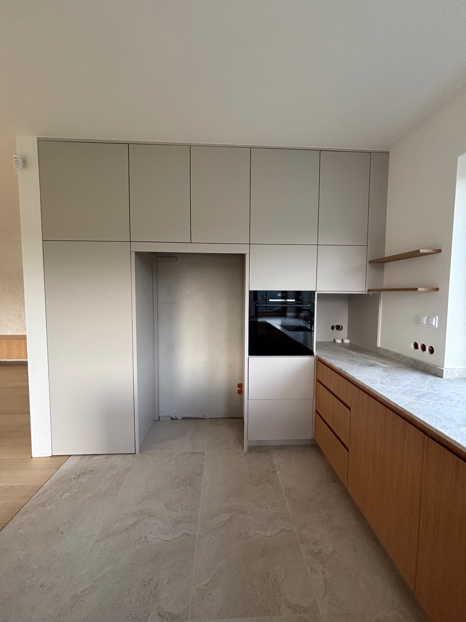 Wnętrze: zabudowa kuchenna w odcieniach szarości i drewna z blatem w kolorze marmuru. Widoczne półki i sprzęt AGD. Płytki podłogowe imitujące kamień.