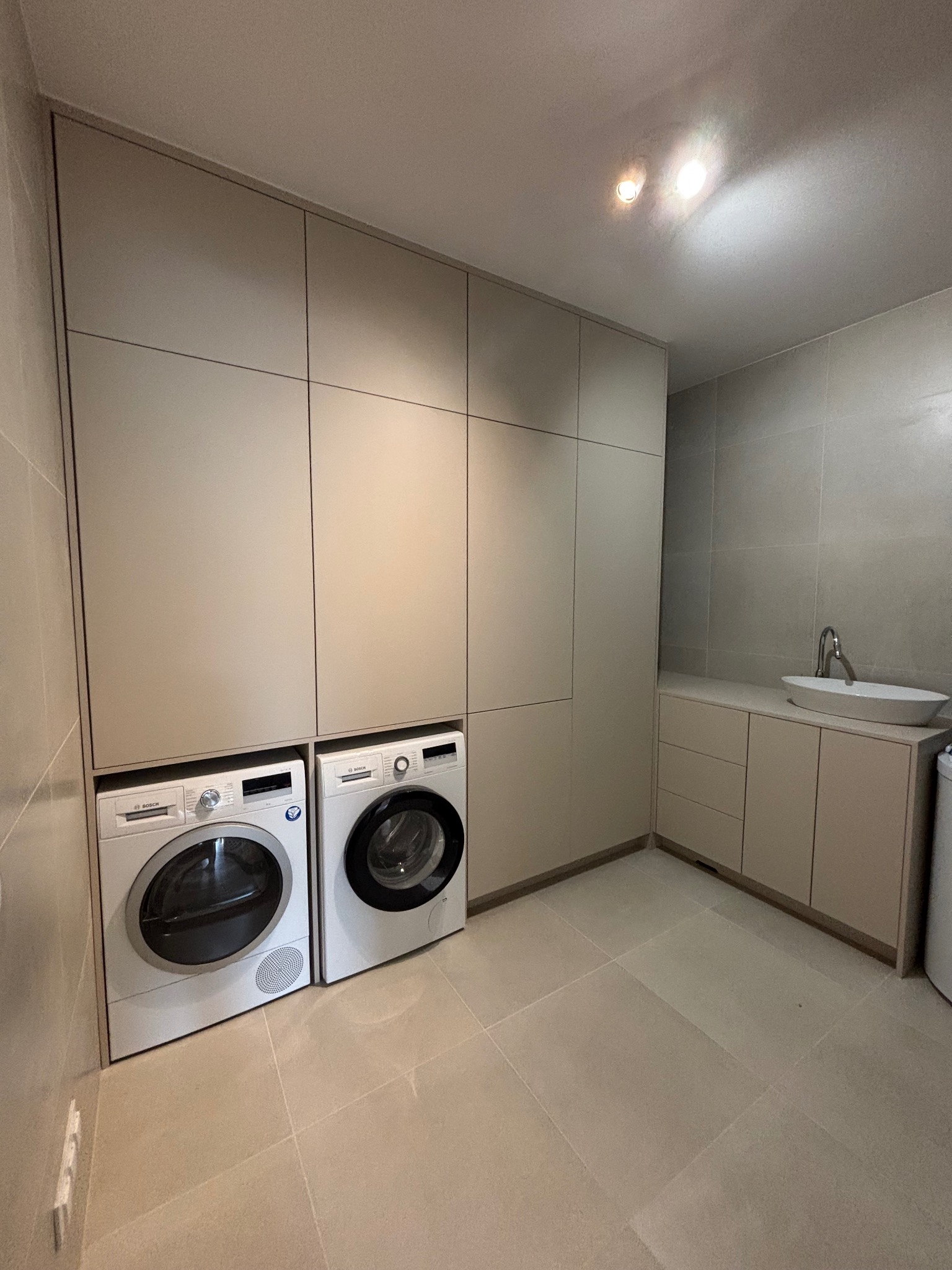 Beżowa zabudowa meblowa z pralką i suszarką Bosch w łazience. Umywalka nablatowa na szafce. Minimalistyczny design, jasne płytki podłogowe i ścienne.