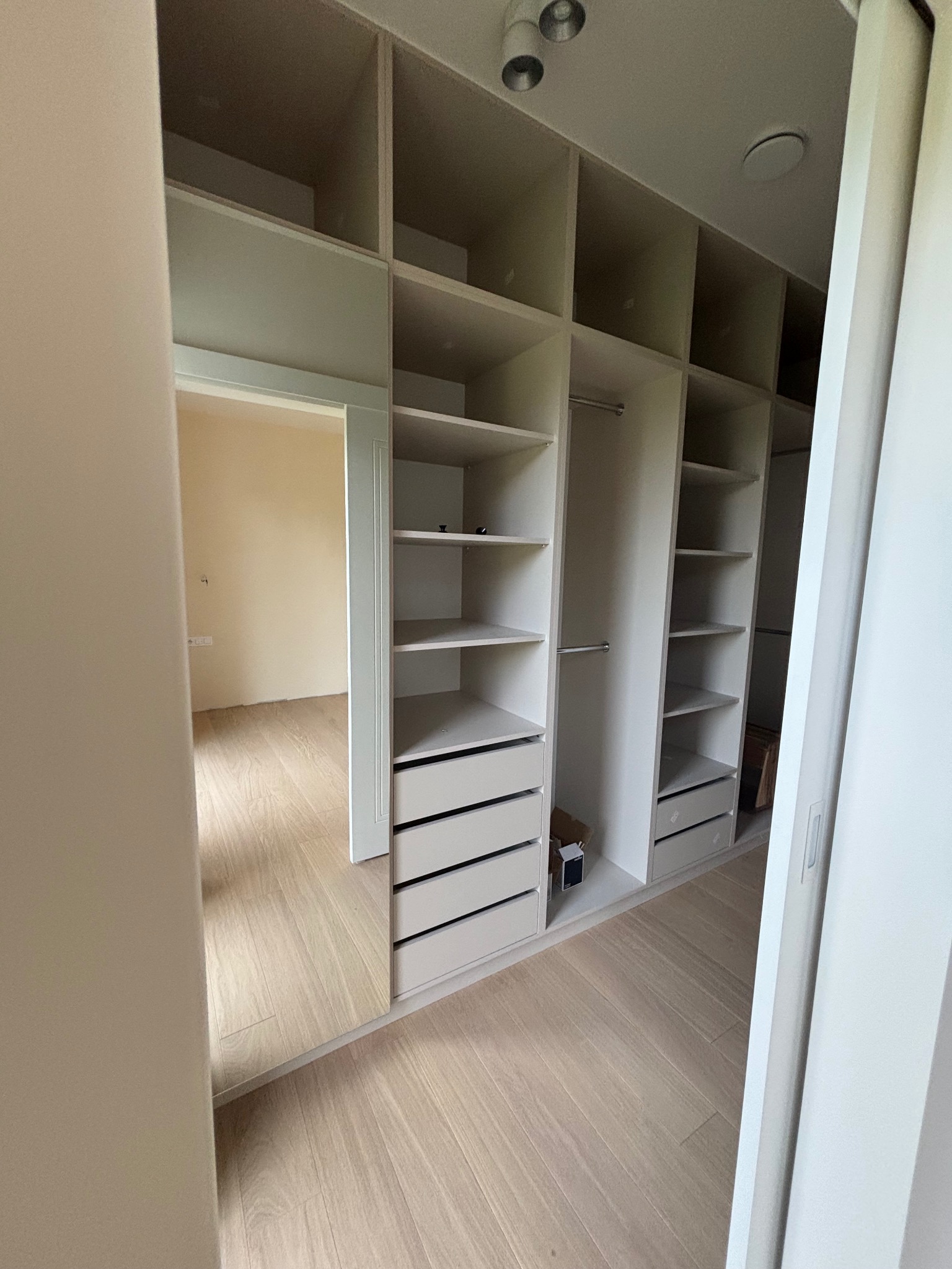 Biała, otwarta garderoba z półkami i szufladami, widok z wnętrza pokoju. Minimalistyczny design, jasne drewno na podłodze, punktowe oświetlenie sufitowe.
