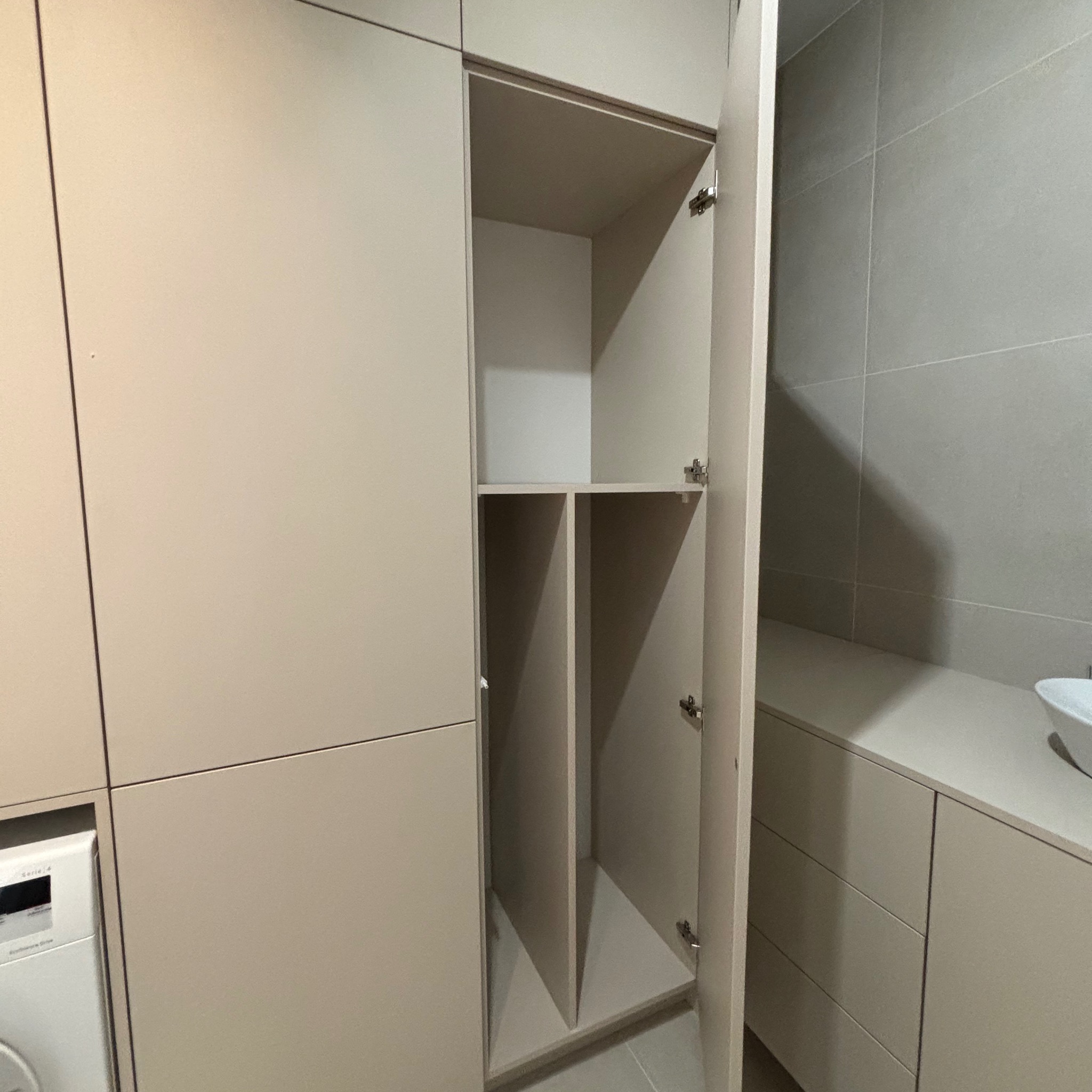 Jasna zabudowa meblowa z otwartą szafką, widoczne półki i przegródki. W tle szare płytki. Obok pralka i umywalka. Minimalistyczny design.