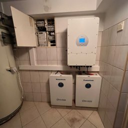 Ciepła Energia - Instalacja magazynów energii w pomieszczeniu z płytkami: dwa białe moduły PowerGem firmy Finaess, biały falownik Deye z wyświetlaczem, skrzynka elektryczna z licznikiem.