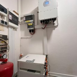 Ciepła Energia - Trzy moduły magazynu energii ułożone jeden na drugim, z podłączonym inwerterem i zabezpieczeniami elektrycznymi na ścianie w pomieszczeniu technicznym.
