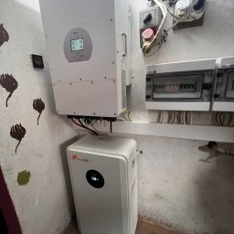 Ciepła Energia - Biały inwerter i magazyn energii Felicity ESS zamontowane na ścianie w pomieszczeniu z widocznymi przewodami i elementami instalacji elektrycznej.