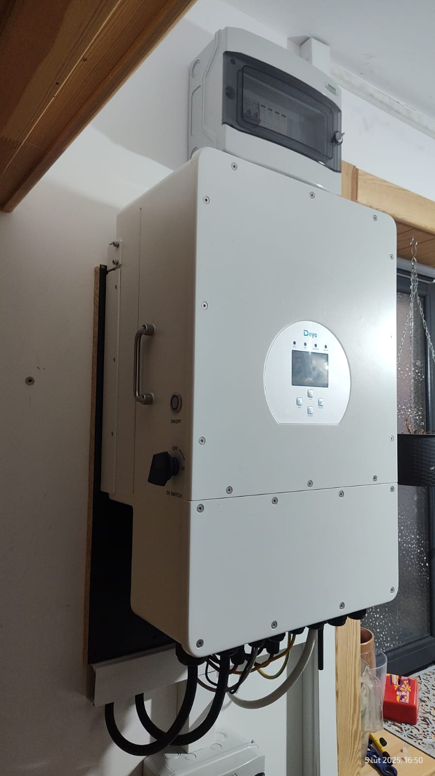 Biały falownik Deye zamontowany na ścianie z widocznymi kablami, wyłącznikiem DC i panelem sterowania, nad nim skrzynka zabezpieczeń.