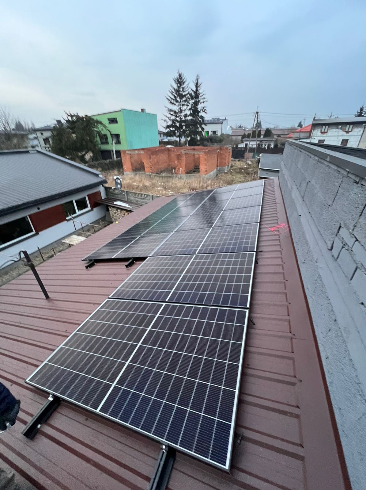Panele słoneczne zamontowane na dachu z blachy trapezowej w pochmurny dzień, widok na okoliczne budynki i niedokończoną budowlę z cegły.