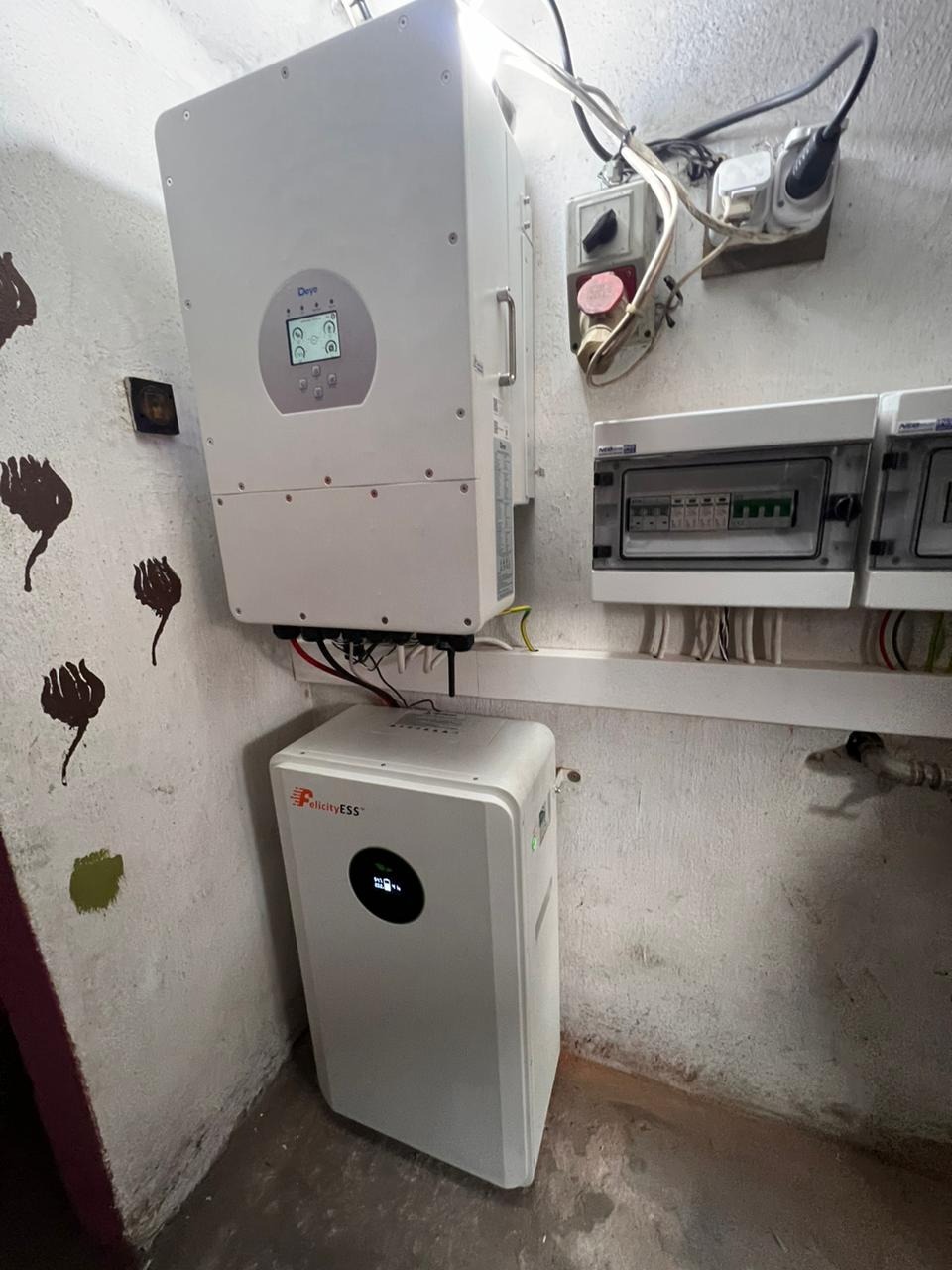 Biały inwerter i magazyn energii Felicity ESS zamontowane na ścianie w pomieszczeniu z widocznymi przewodami i elementami instalacji elektrycznej.