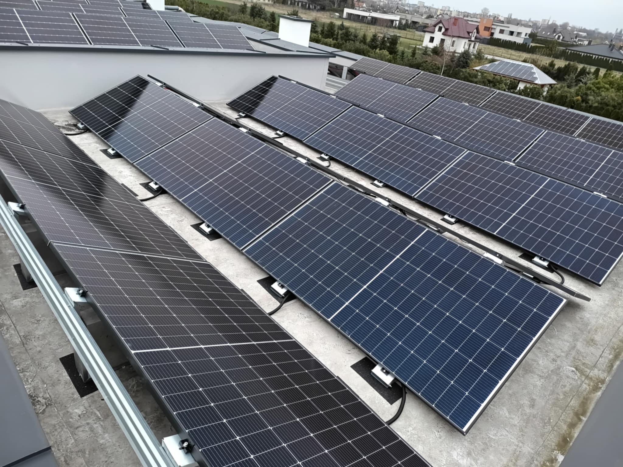 Rząd paneli słonecznych na płaskim dachu budynku mieszkalnego, widoczne elementy montażowe i przewody, w tle zabudowa jednorodzinna.