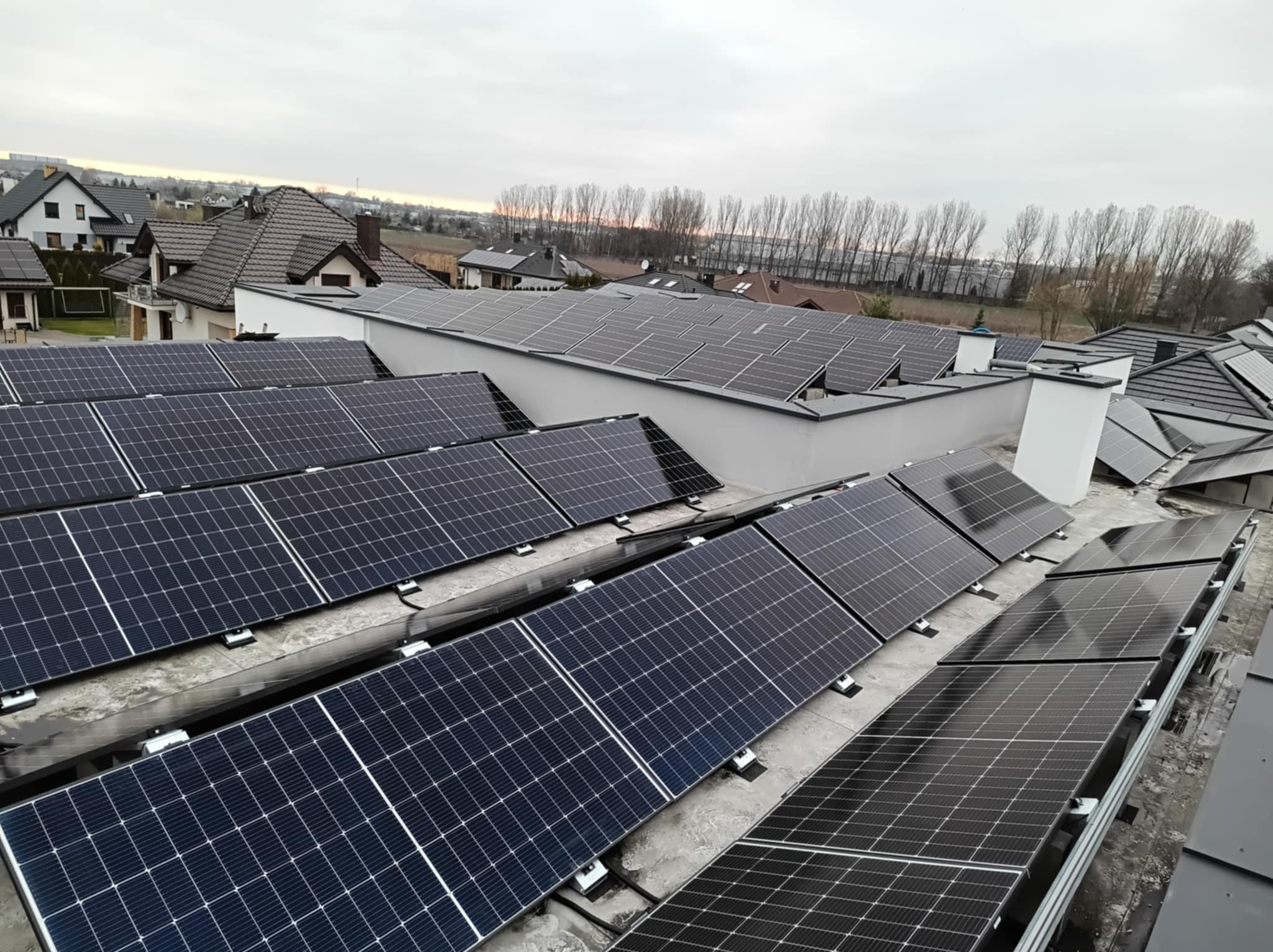 Widok z góry na instalację paneli słonecznych na dachu budynku mieszkalnego, z widocznymi elementami montażowymi i otaczającą zabudową w tle.