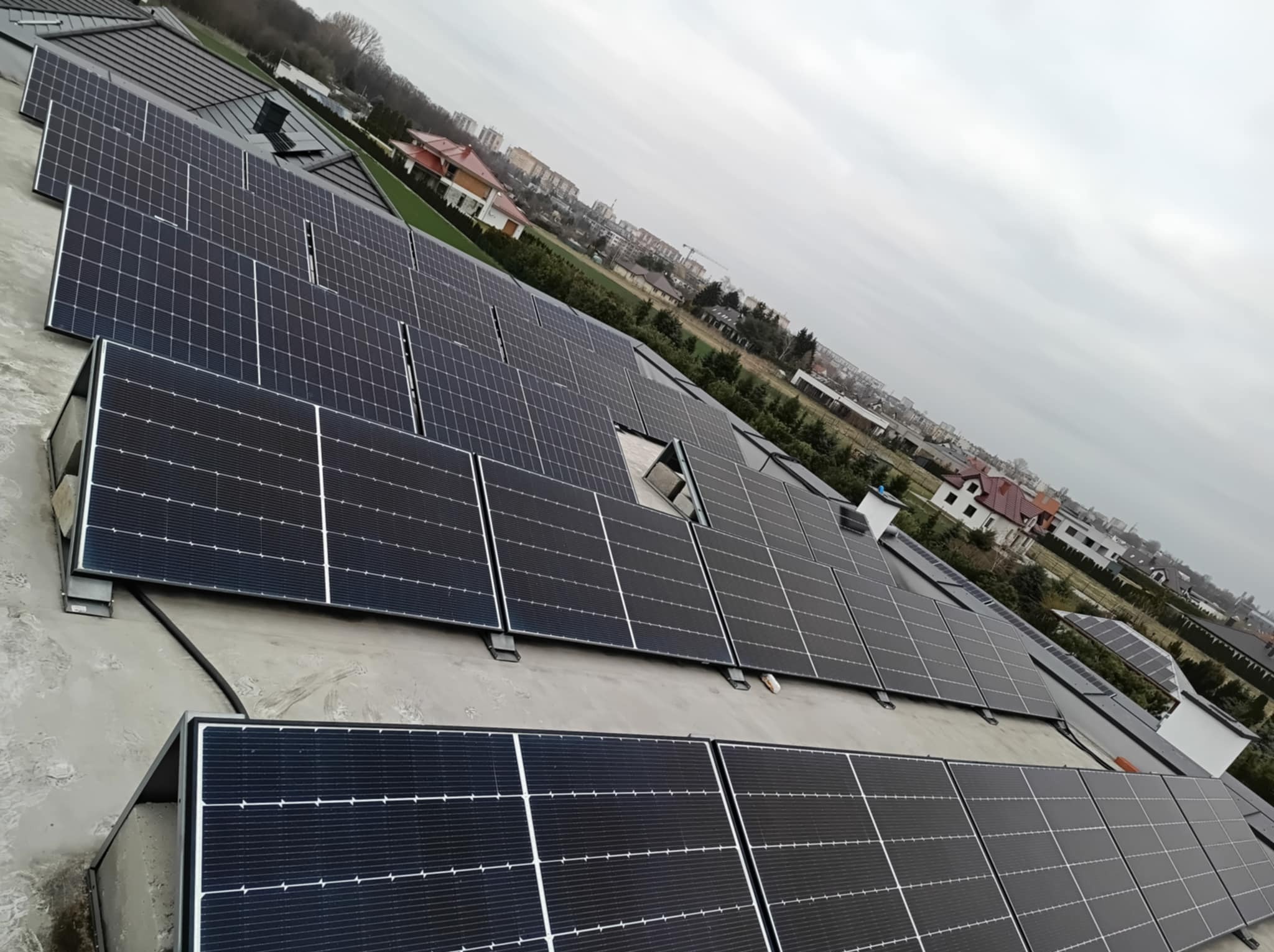 Instalacja paneli słonecznych na płaskim dachu w Radomiu, widoczne panele z czarnymi ramami, szare niebo w tle, perspektywa ukośna.