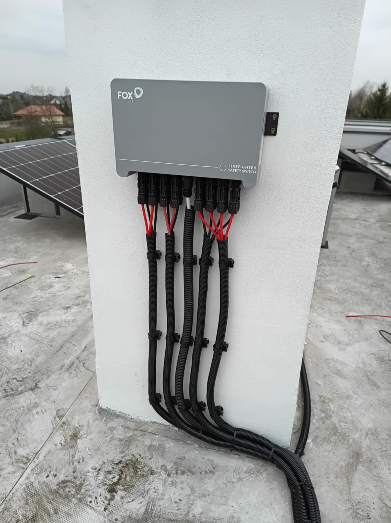 Szary moduł z napisem FOX ESS i FIRE FIGHTER SAFETY SWITCH zamontowany na białym murze, z którego wychodzą czarne rurki instalacyjne z czerwonymi kablami, widoczne panele na dachu i domy w tle.