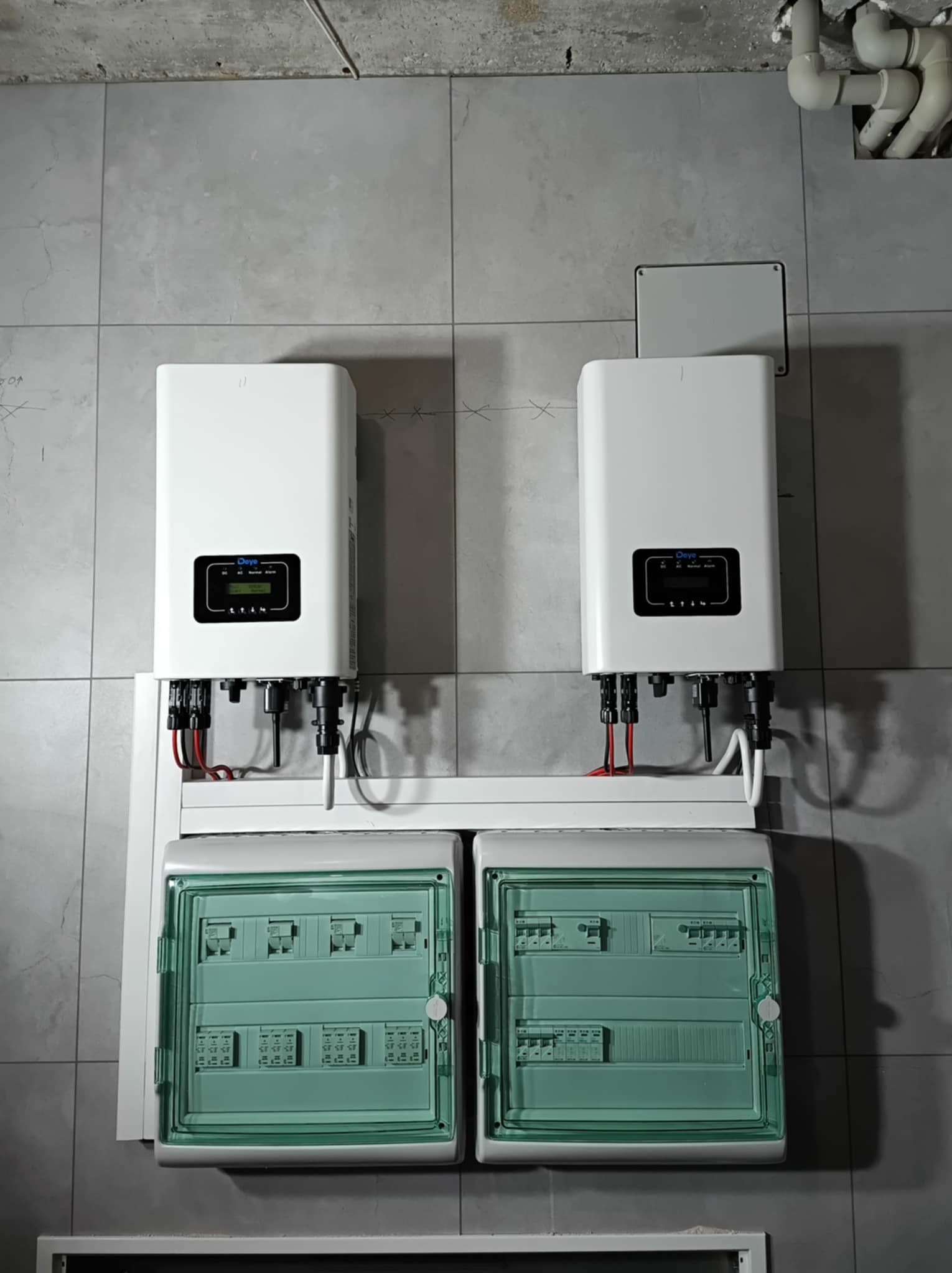Dwa białe falowniki marki Deye zamontowane na ścianie z szarych płytek, poniżej dwie zielone skrzynki rozdzielcze.