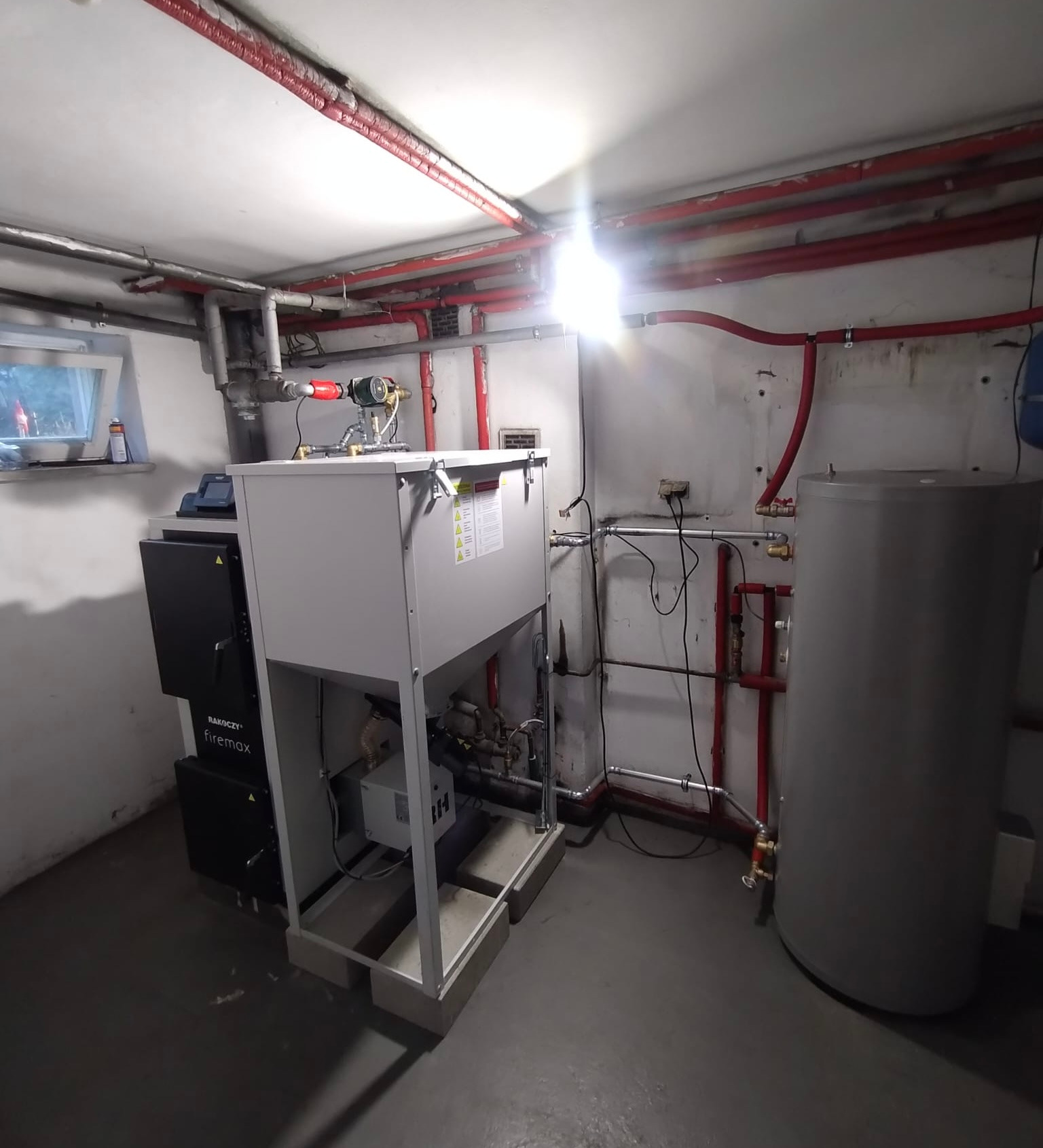 Zainstalowany piec CO marki Rakoczy Firemax w kotłowni z widocznymi rurami instalacji grzewczej i zasobnikiem ciepłej wody.
