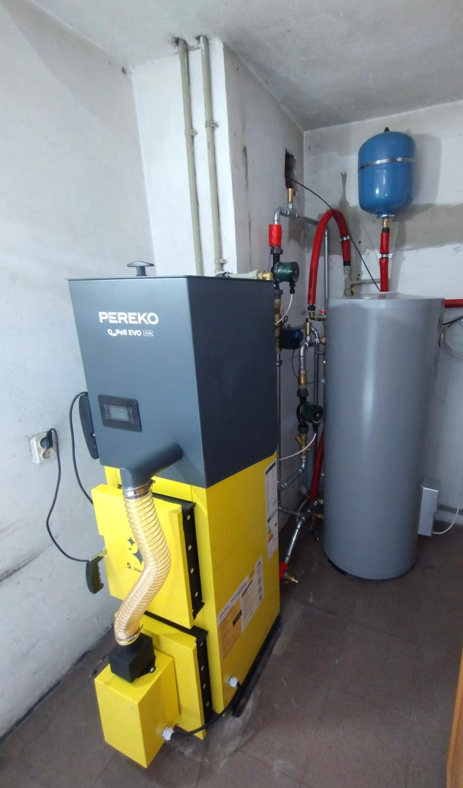 Żółto-szary piec Pereko Q-Pell EVO 15kW z podłączonymi instalacjami w kotłowni, obok szary zbiornik wyrównawczy i niebieski zbiornik ciśnieniowy.