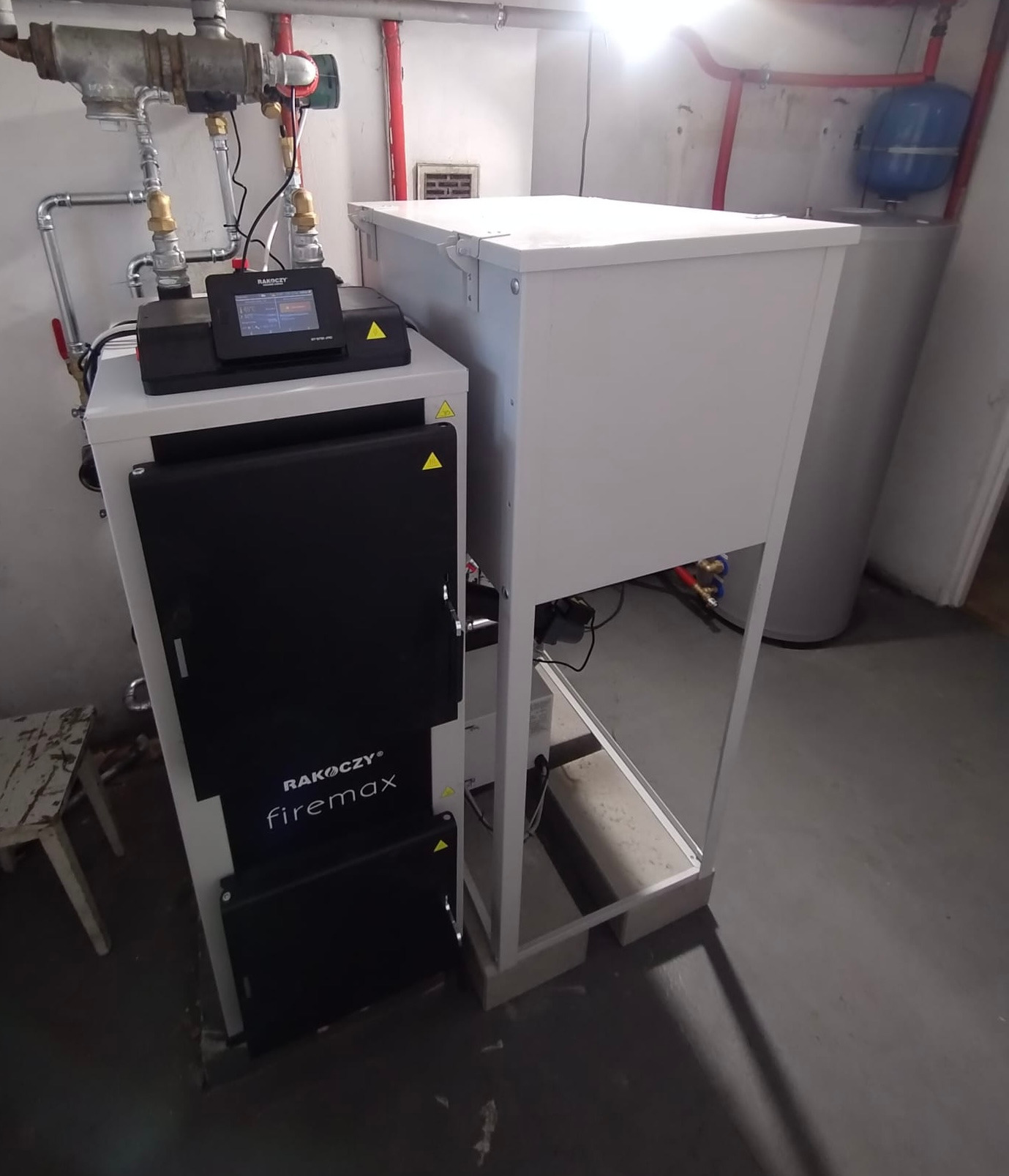 Kocioł RAKOCZY firemax z podajnikiem, z elektronicznym panelem sterowania. Widoczne rury instalacji grzewczej i zasobnik wody. Szare tło posadzki.