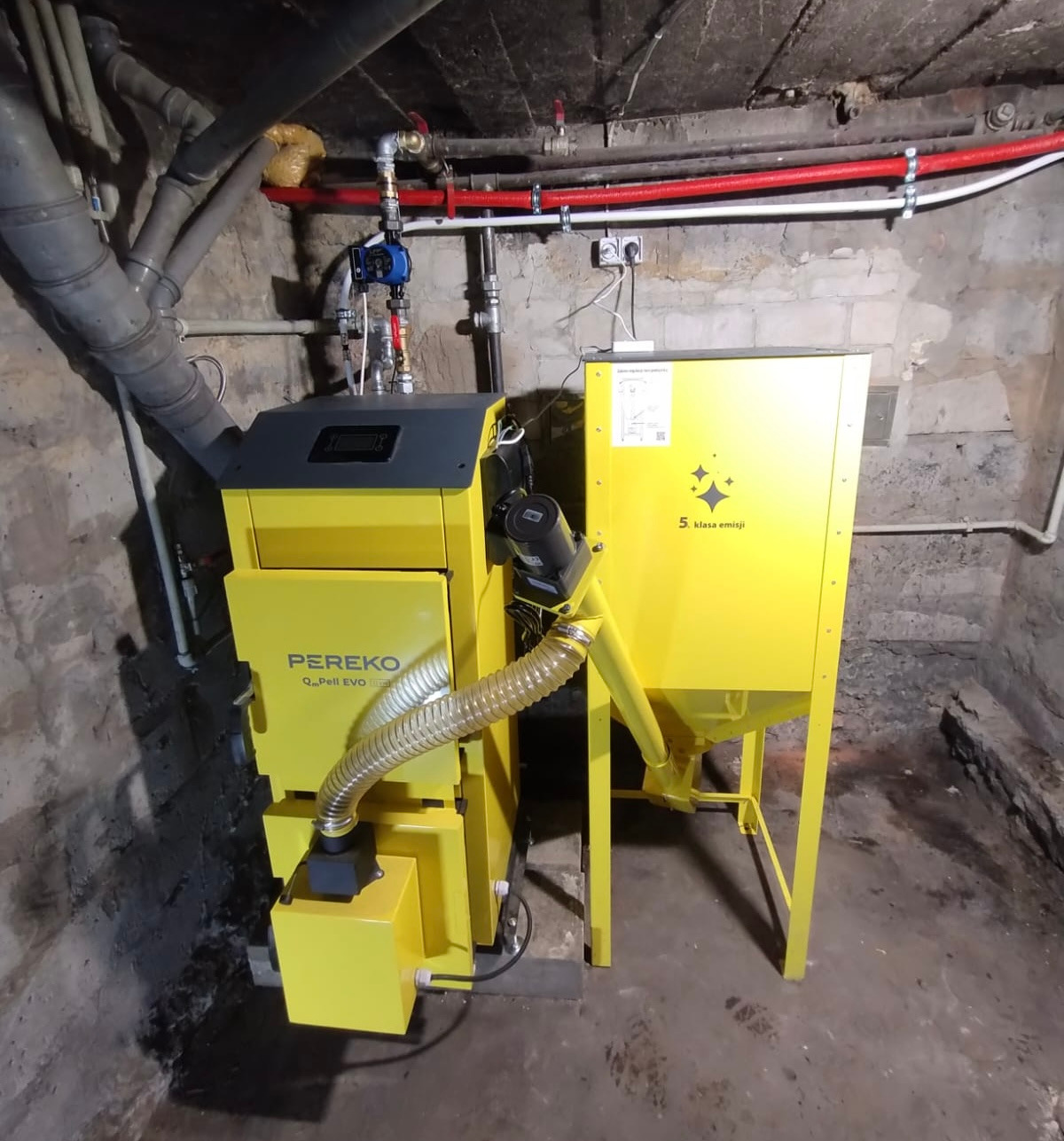 Żółty piec na pellet marki PEREKO w piwnicy, widoczne rury instalacyjne na ścianie. Piec z podajnikiem, klasa emisji 5.