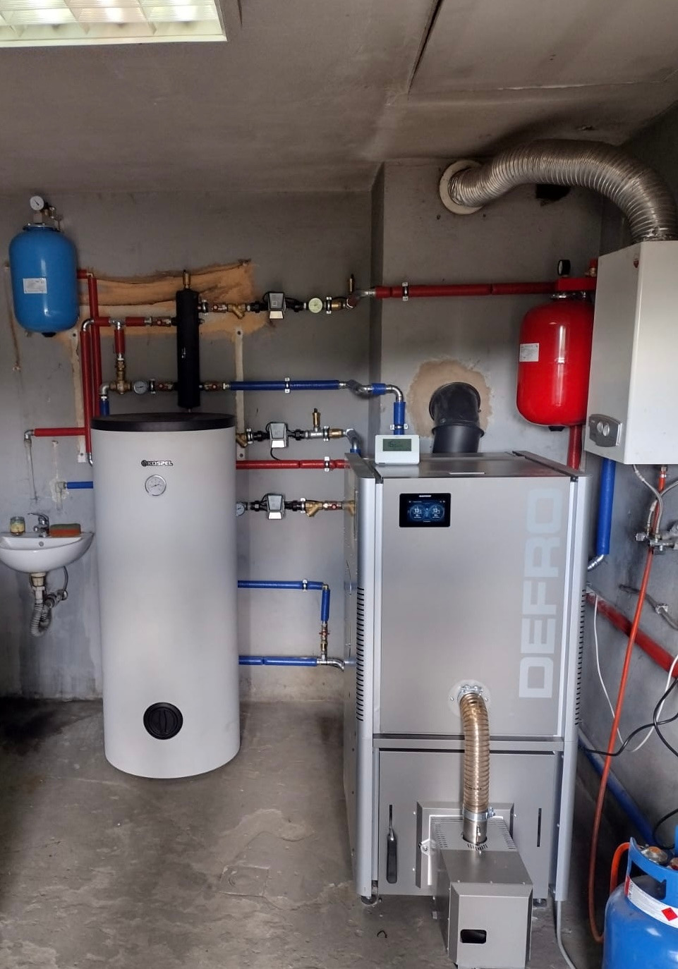 Zainstalowany piec CO Defro obok zbiornika Kospel, z widoczną instalacją hydrauliczną (czerwone i niebieskie rury) w kotłowni. Umywalka z kranem po lewej.