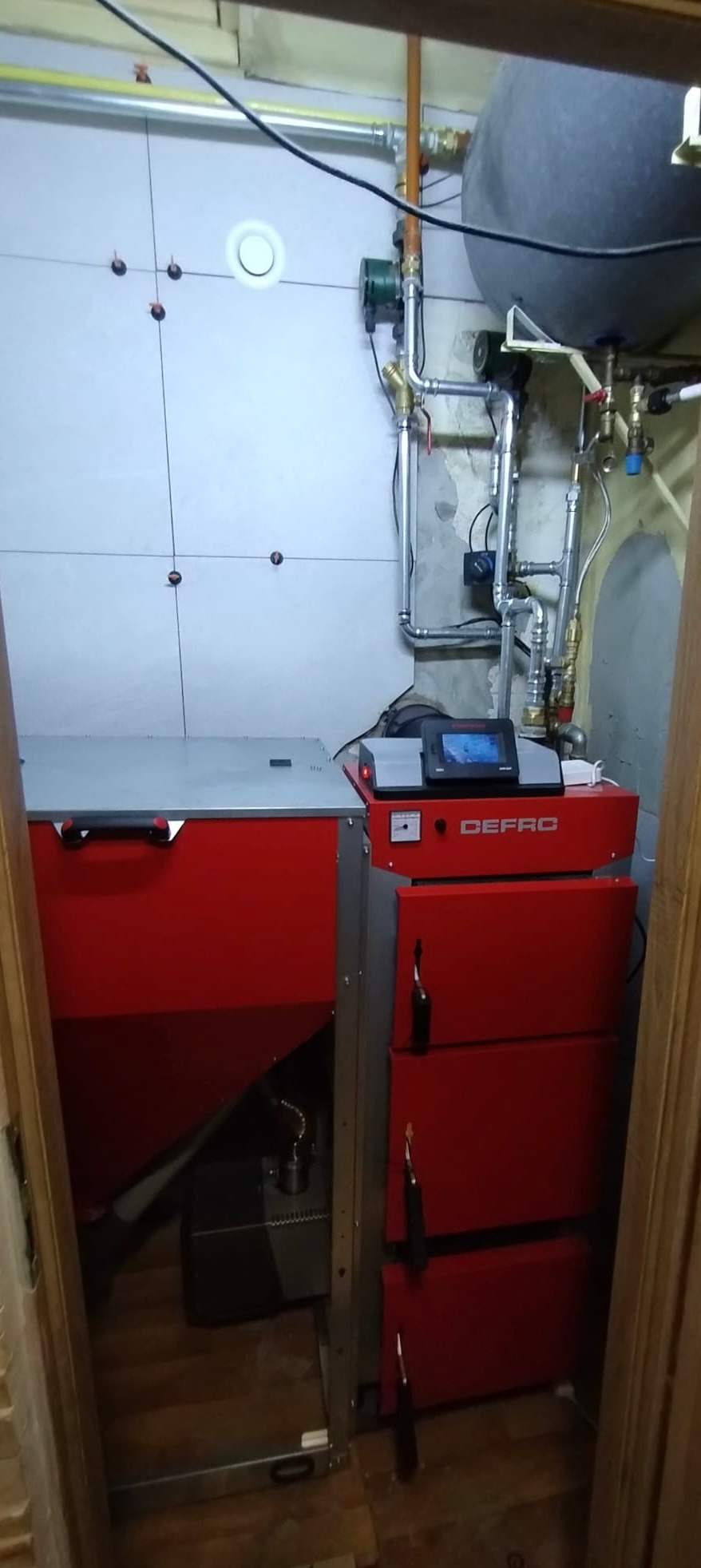 Czerwony piec CO marki Defro w kotłowni, widoczne rury instalacji grzewczej i zasobnik. Na ścianie białe płytki.