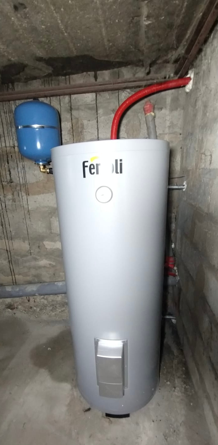 Bojler Ferroli w piwnicy z instalacją: czerwona rura, niebieski zbiornik wyrównawczy. Surowy beton na ścianach i suficie. Szary betonowy podkład.