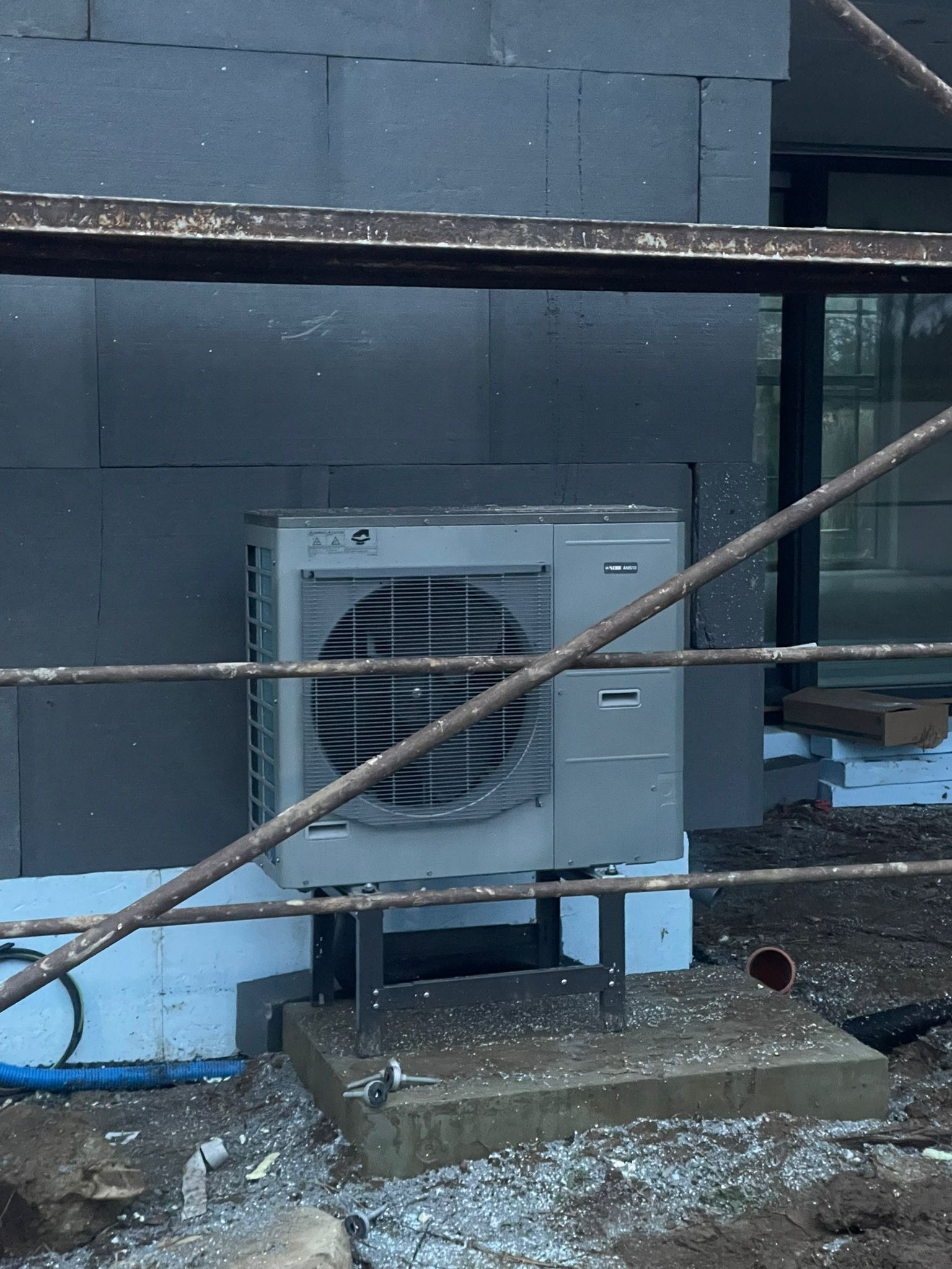 Zamontowana zewnętrzna jednostka pompy ciepła na fundamencie, widoczna przez rusztowanie na tle ocieplonej ściany budynku. Elementy instalacji na ziemi.