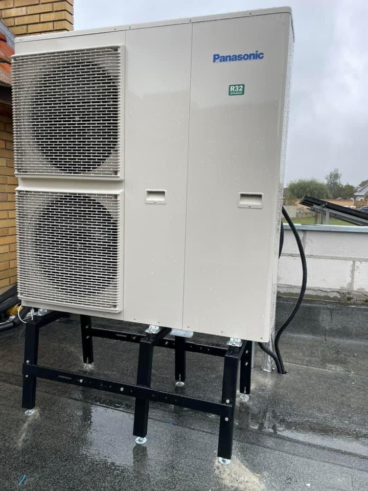 Zamontowana zewnętrzna jednostka pompy ciepła Panasonic R32 na dachu, na czarnej, metalowej konstrukcji. Widoczny fragment budynku z cegły i mokra nawierzchnia.