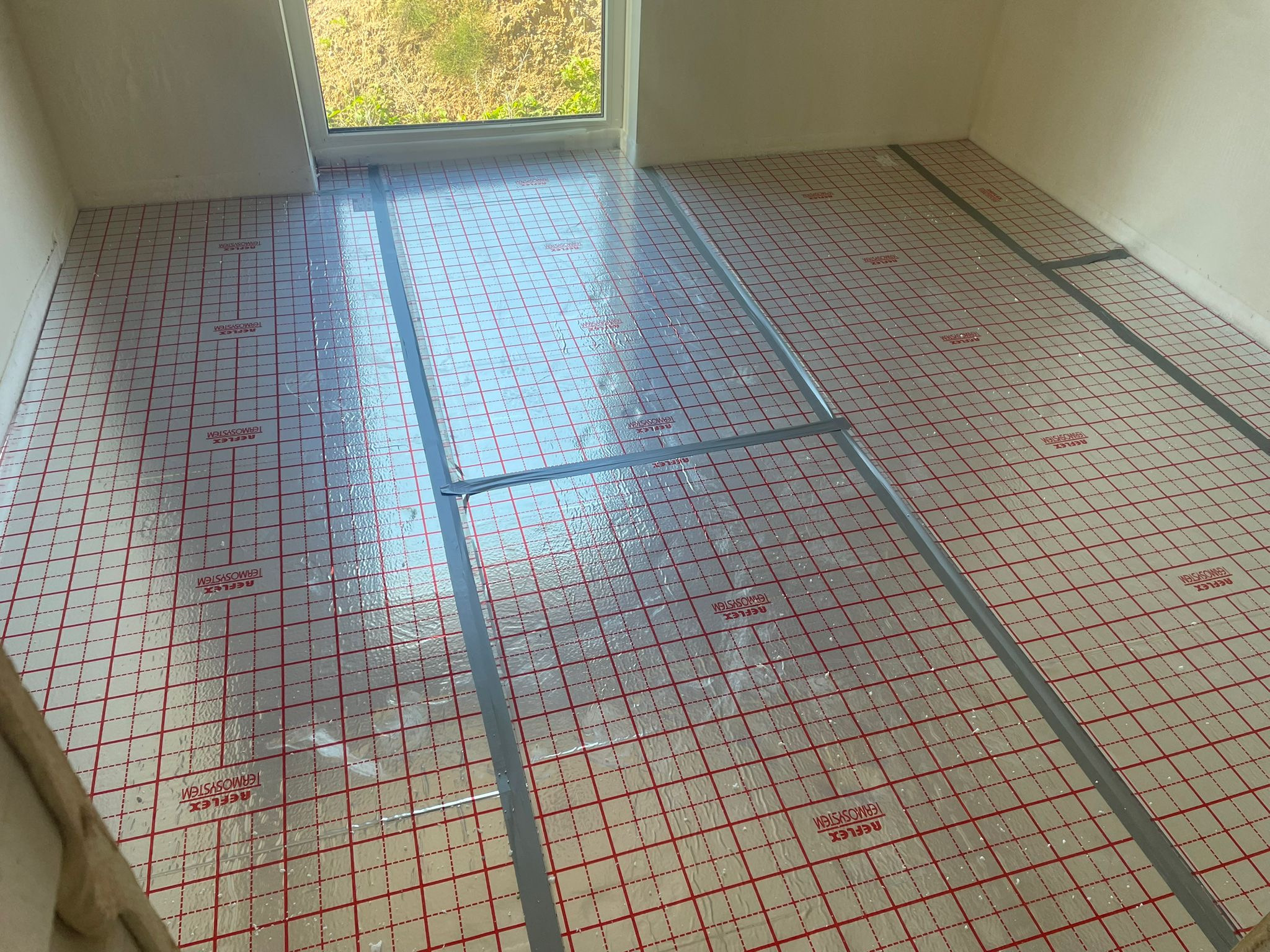 Przygotowanie do montażu ogrzewania podłogowego: rozłożona mata izolacyjna Reflex Termosystem z czerwoną siatką i srebrną folią, zabezpieczona taśmą. Widok z góry na wnętrze.