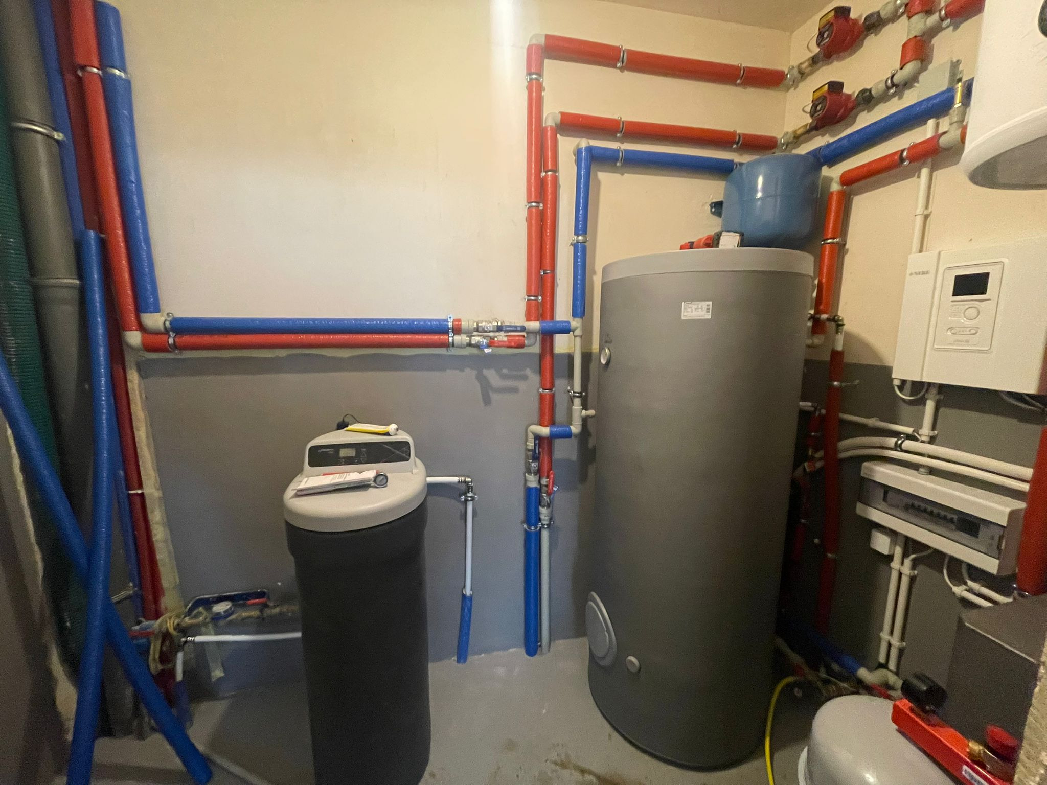Szara instalacja kotłowni z rurami w kolorze czerwonym i niebieskim, zbiornikiem na wodę i zmiękczaczem wody. Widoczne elementy montażowe i sterownik.