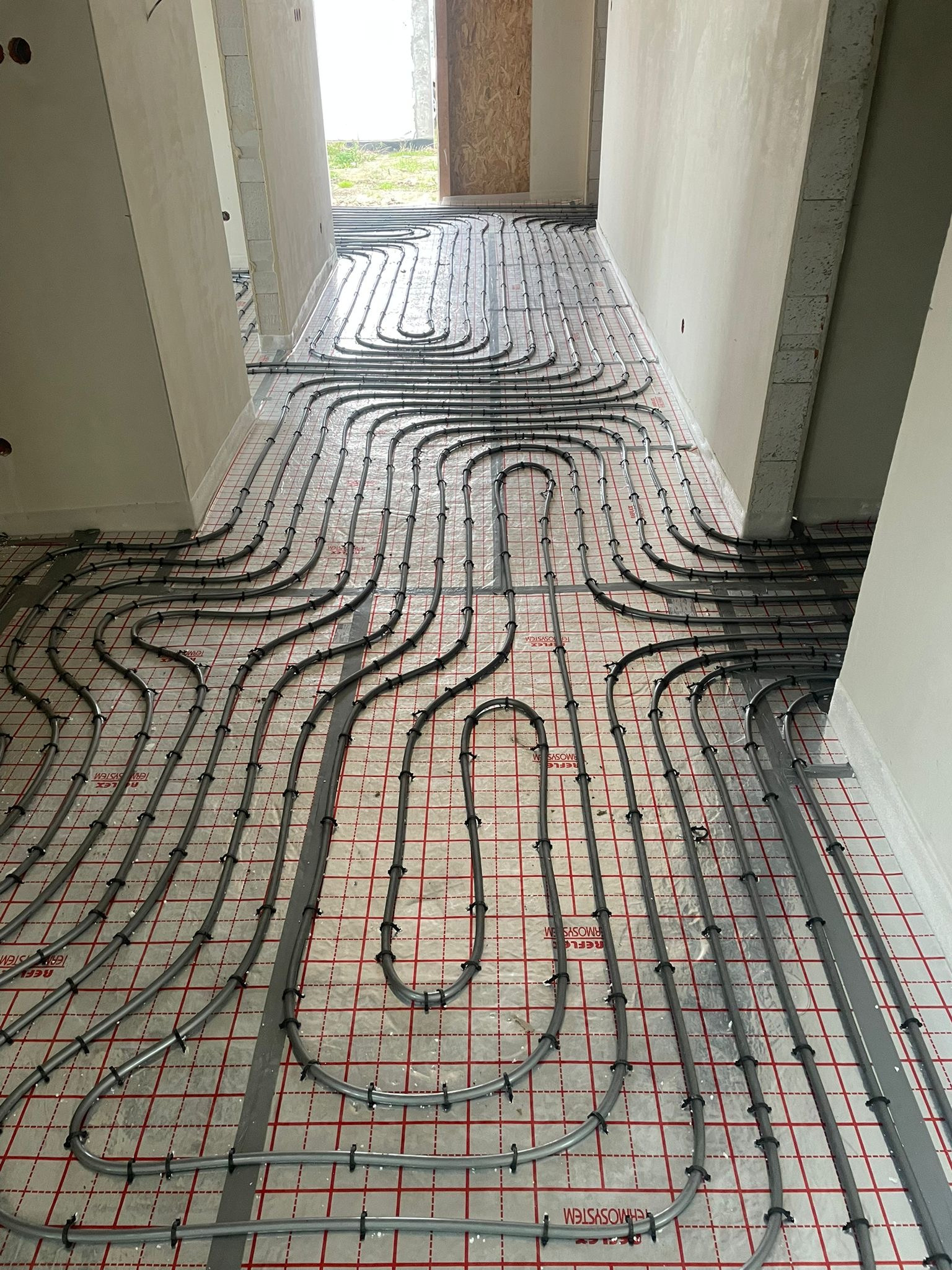 Szare rury ogrzewania podłogowego ułożone w pętle na folii z czerwoną siatką, widok z góry w surowym wnętrzu budynku w trakcie budowy.