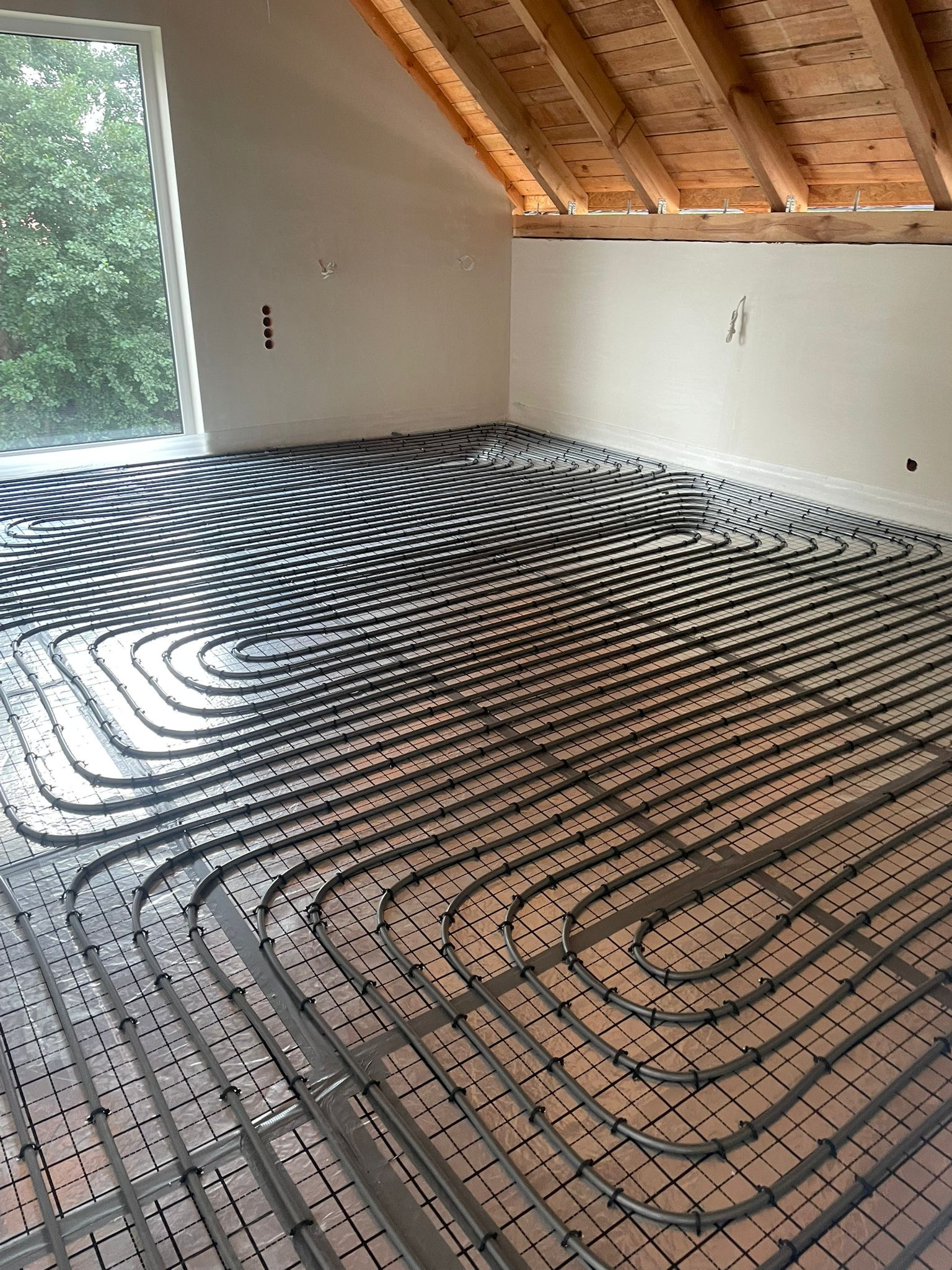 Instalacja ogrzewania podłogowego w nowym domu. Równoległe rury grzewcze ułożone na folii izolacyjnej, widoczne drewniane elementy konstrukcji dachu i okno.