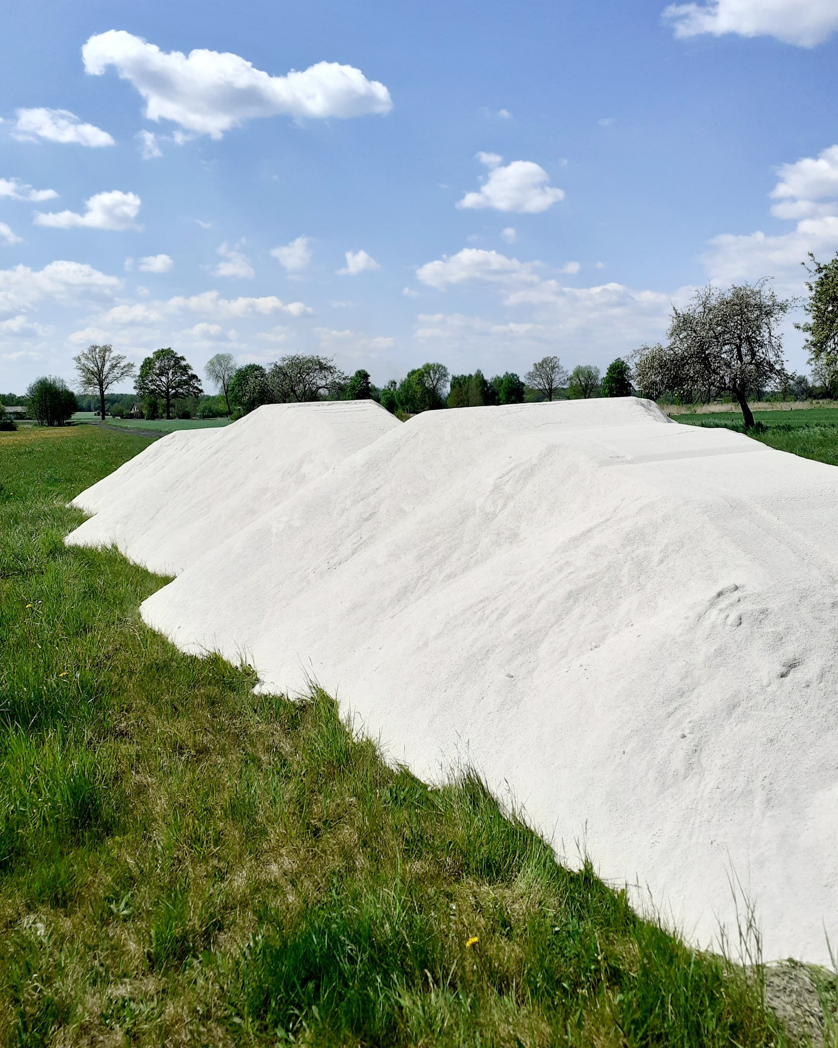 Duża pryzma białego materiału sypkiego, prawdopodobnie wapna lub piasku, na zielonej łące pod błękitnym niebem z białymi chmurami.