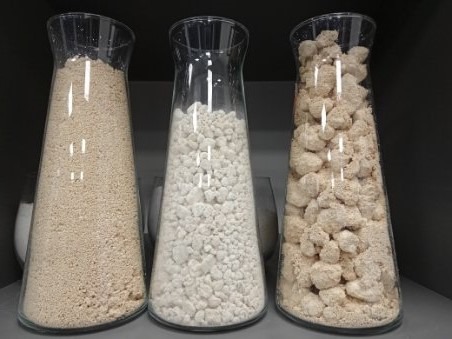 Trzy szklane wazony o stożkowym kształcie wypełnione różnymi materiałami sypkimi: drobny piasek, białe granulki i nieregularne bryłki w odcieniach beżu.