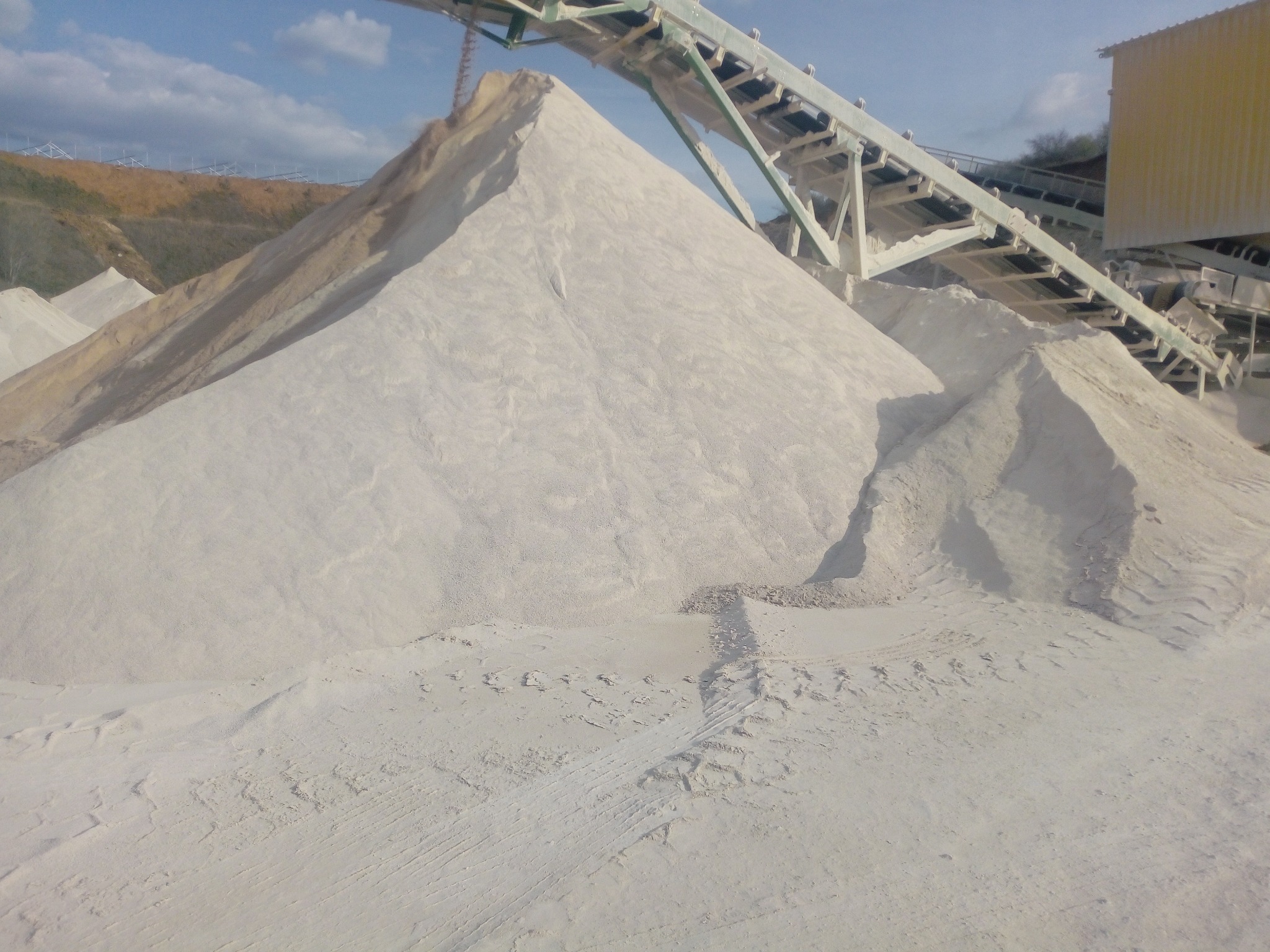 Góra jasnego kruszywa pod taśmociągiem na terenie kopalni, widoczne ślady opon