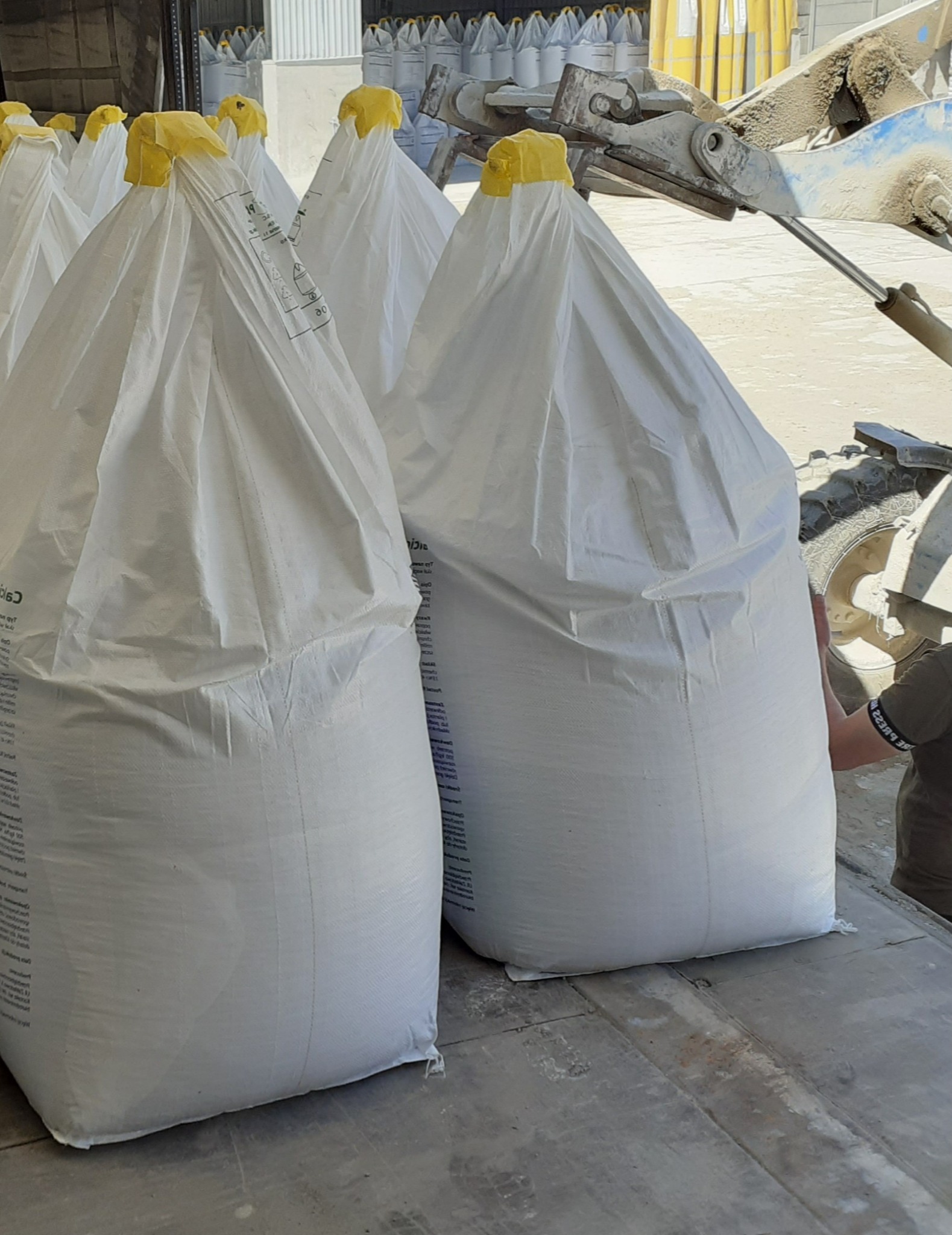 Białe worki typu big bag z żółtymi zamknięciami, załadowane na platformę transportową, w tle widoczne kolejne worki i ramię maszyny załadunkowej.
