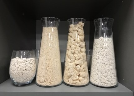 Cztery szklane naczynia różnej wielkości wypełnione jasnymi materiałami sypkimi i bryłowymi o różnej granulacji, ustawione na szarej półce.