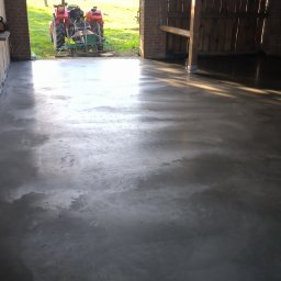 RBBet-Rafał Blacha - Świeżo wylana, gładka posadzka przemysłowa wewnątrz budynku gospodarczego. Na zewnątrz, przez otwarte wrota, widać traktor na zielonym trawniku.