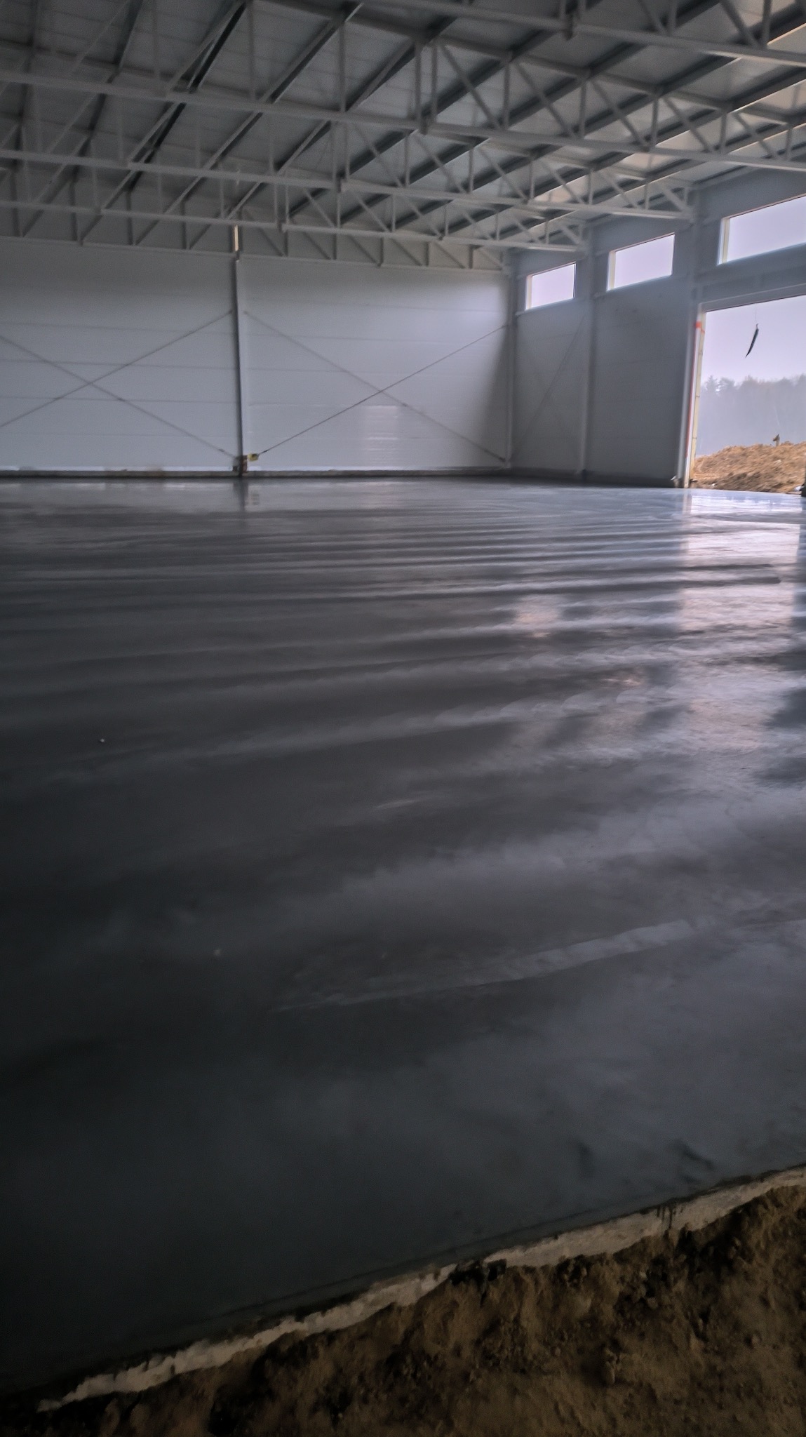 Świeżo wylana, mokra, szara posadzka w hali przemysłowej z widoczną konstrukcją dachu i bramą wjazdową.