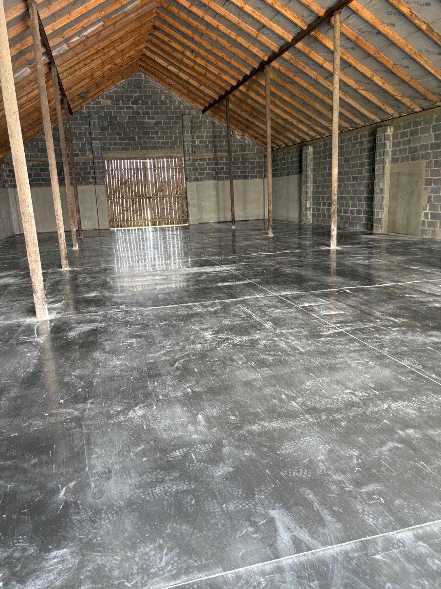 Gładka, szara posadzka przemysłowa w surowym wnętrzu z drewnianą konstrukcją dachu i szarą ścianą z pustaków. Widoczne ślady butów na posadzce.