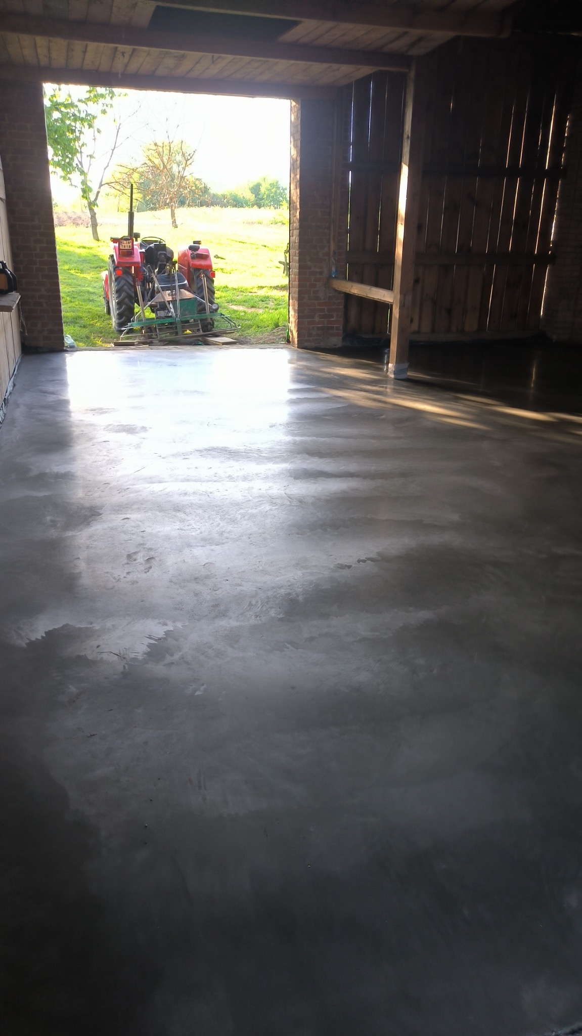 Świeżo wylana, gładka posadzka przemysłowa wewnątrz budynku gospodarczego. Na zewnątrz, przez otwarte wrota, widać traktor na zielonym trawniku.