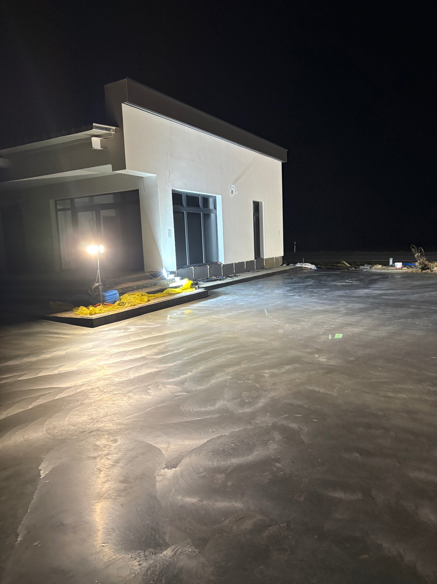 Wylana, szara posadzka przemysłowa w nowym budynku, oświetlona lampą budowlaną. Widoczne ślady po zacieraniu. Prace w toku, nocne oświetlenie.