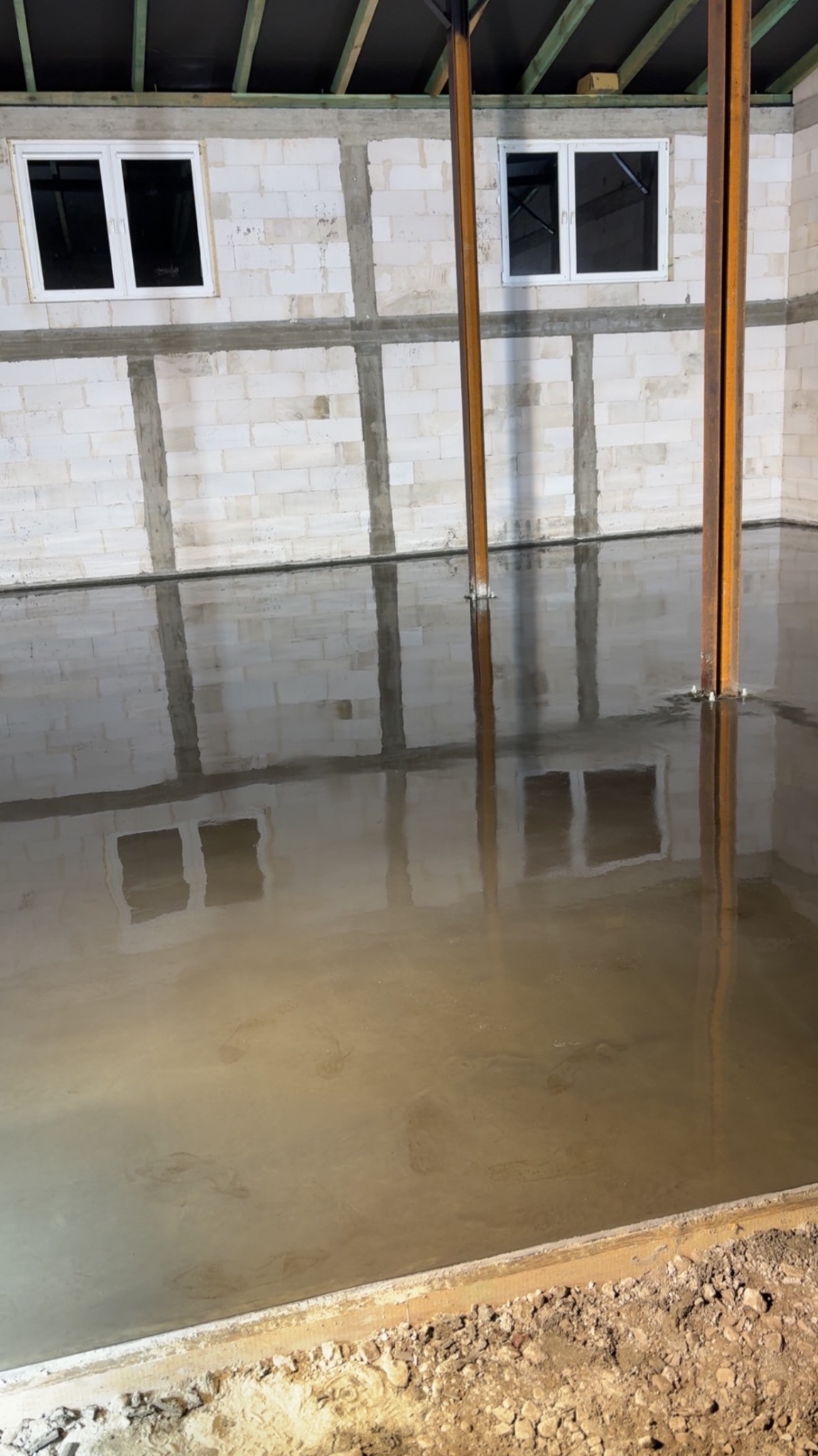 Świeżo wylana, gładka posadzka przemysłowa w surowym wnętrzu z oknami. Widoczne metalowe podpory i ściany z bloczków betonowych. Podłoże przygotowane pod dalsze prace.