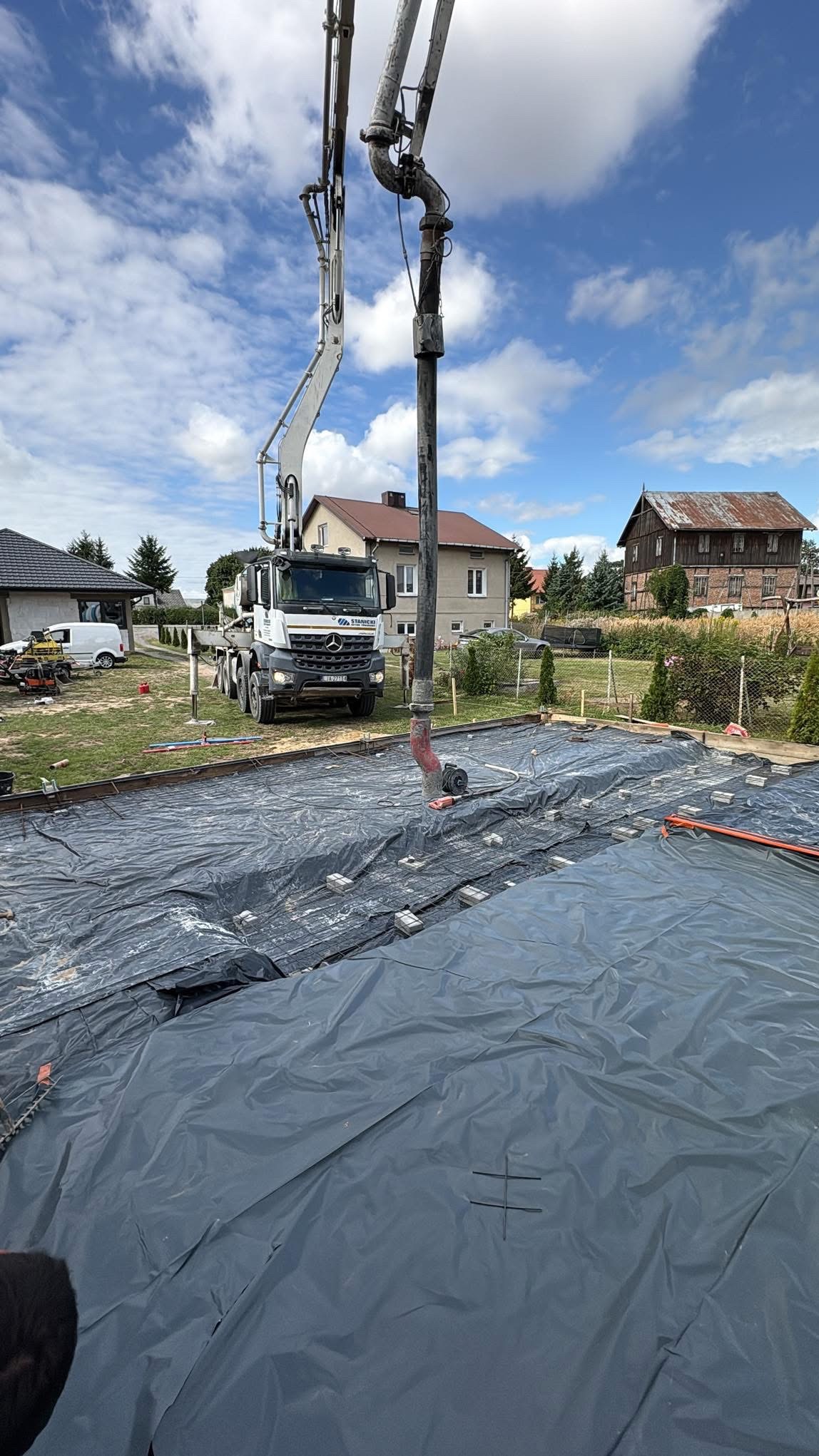 Wylewka betonu na folii budowlanej z użyciem pompy do betonu na tle domów jednorodzinnych i błękitnego nieba z chmurami. Widoczne zbrojenia i szalunki.
